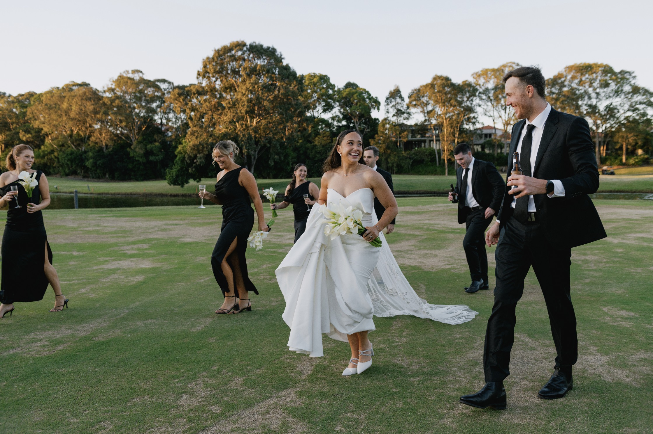 A bride and friends celebrating on a golf course during sunset, with the bride in a white gown holding a bouquet and others dressed in black, all smiling and holding drinks.