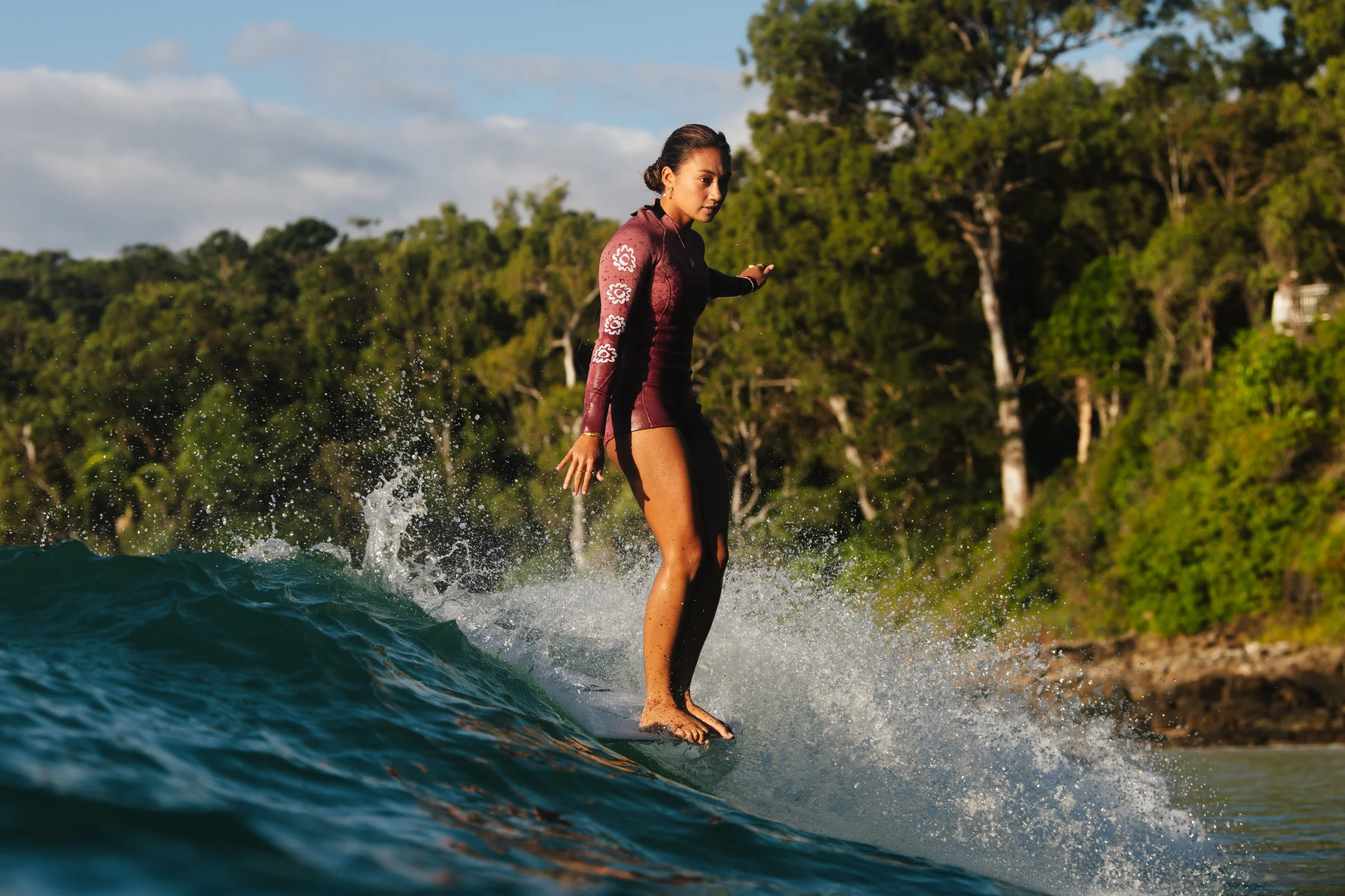 Lilliana Bowrey standing on a surfboard riding a small wave near the shoreline with trees in the background.