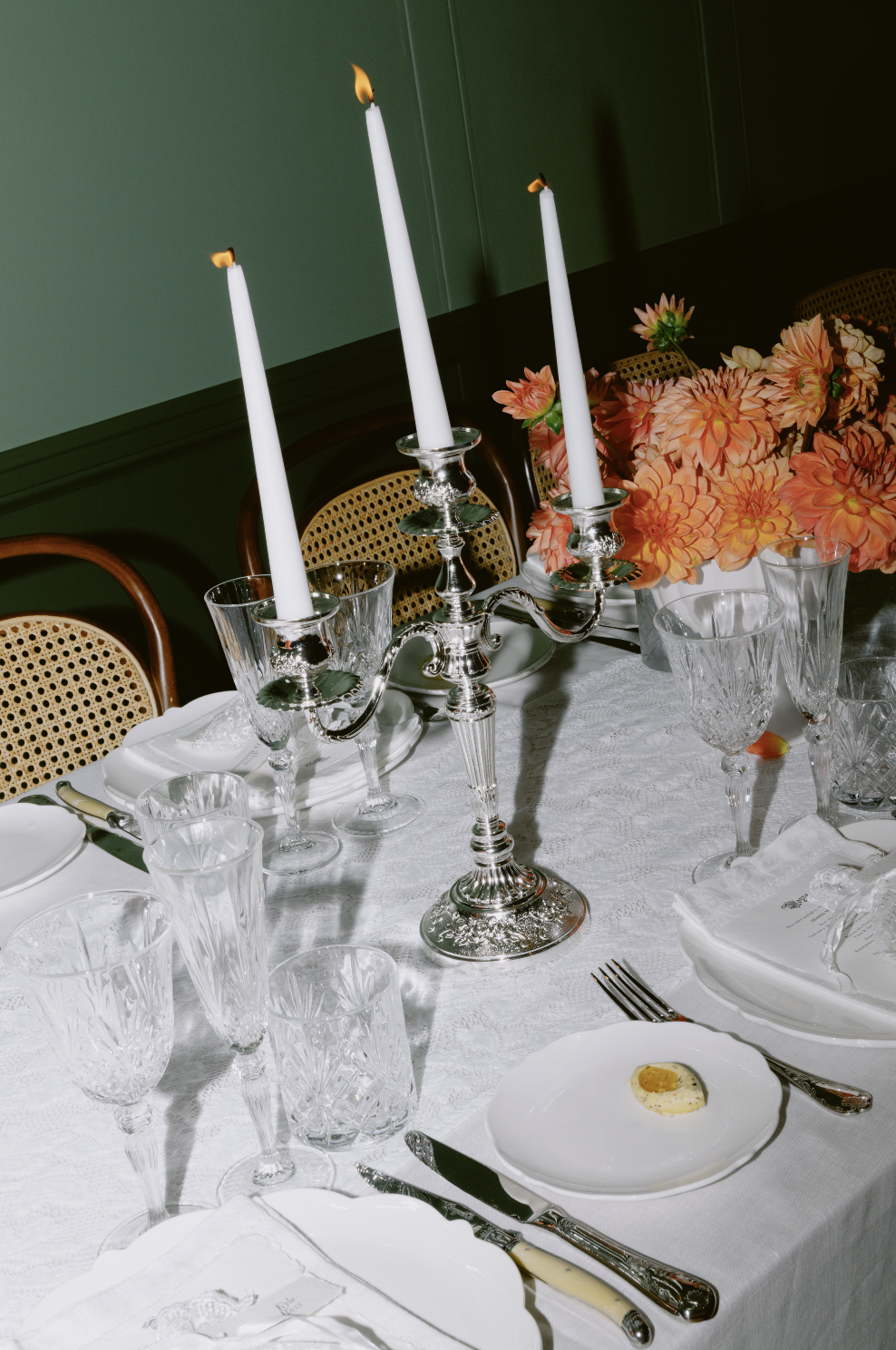 A decorated dining table with a silver candelabrum holding three lit white candles, multiple glasses, white plates, silver cutlery, a floral centerpiece with pink flowers, and white tablecloth.
