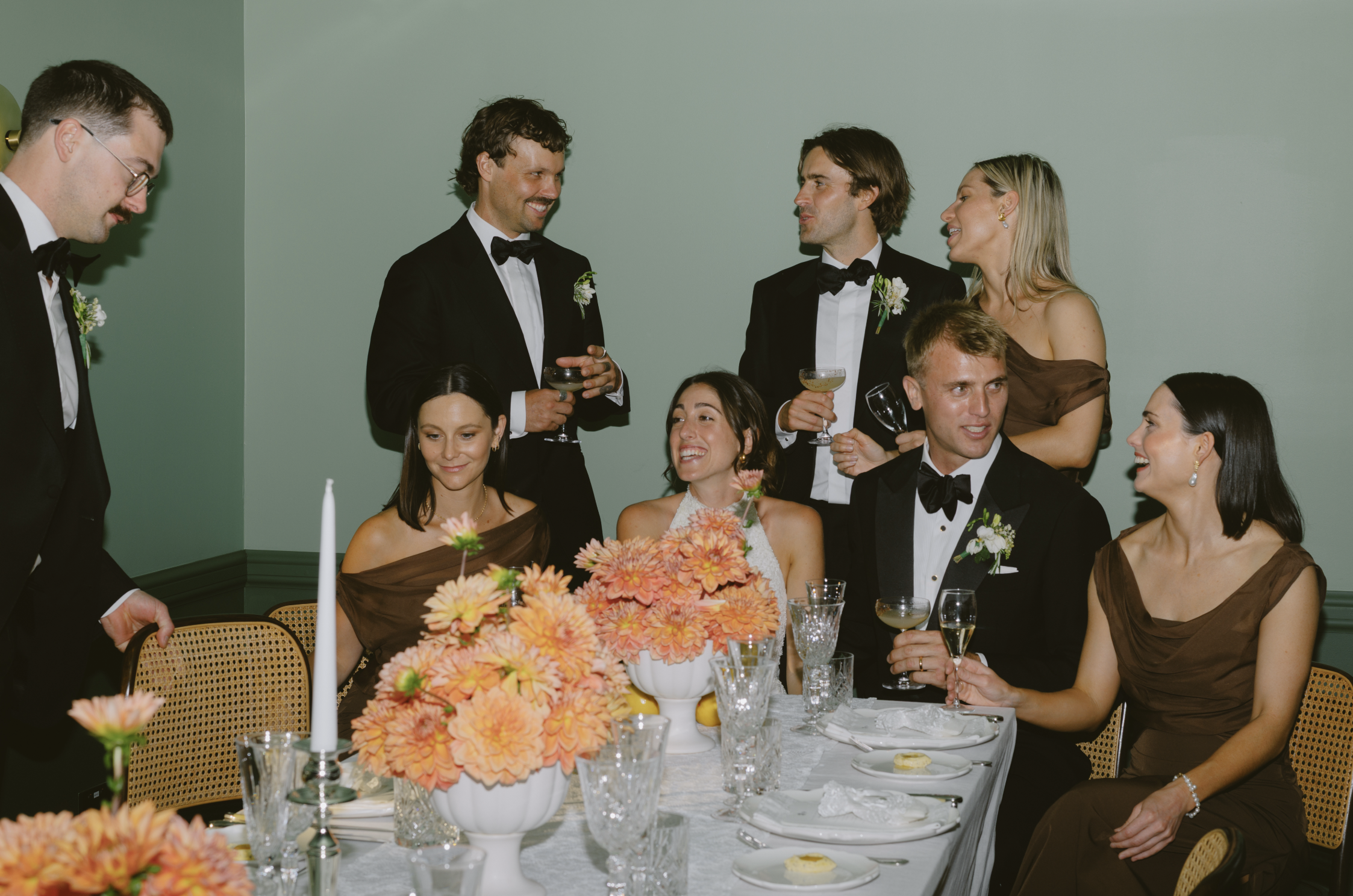 People dressed in formal attire gathered around a dinner table at a celebration or wedding reception, with floral centerpieces, candles, and glassware.