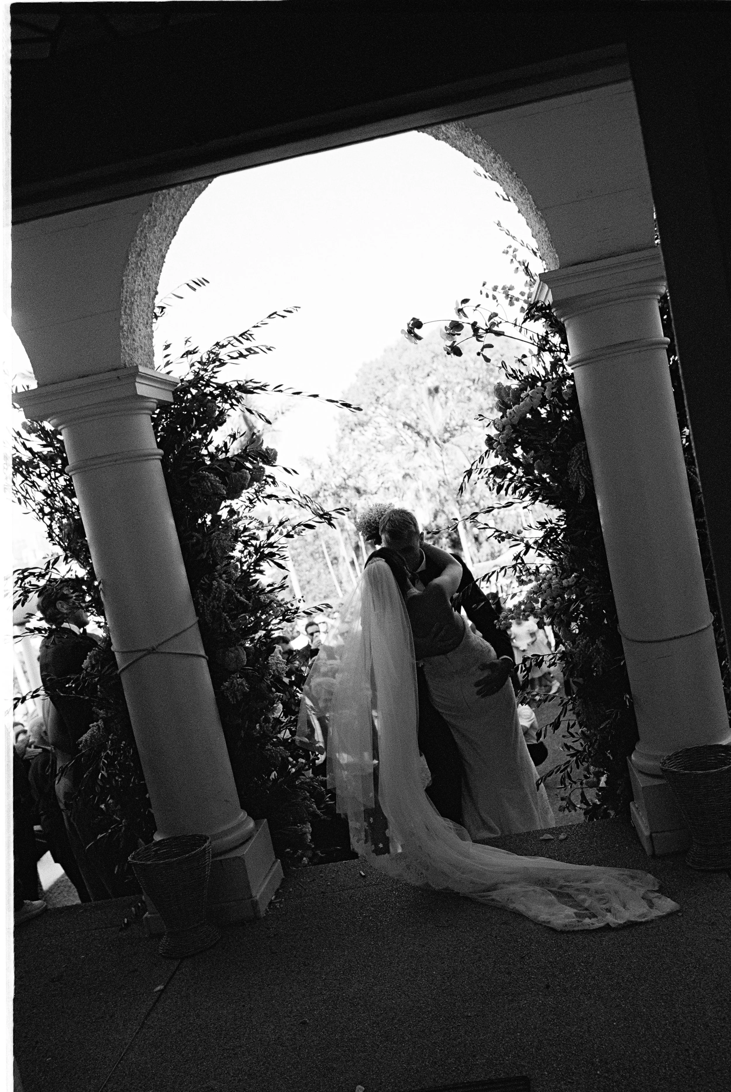 A wedding couple kissing under a decorative archway with floral arrangements, with wedding guests in the background.