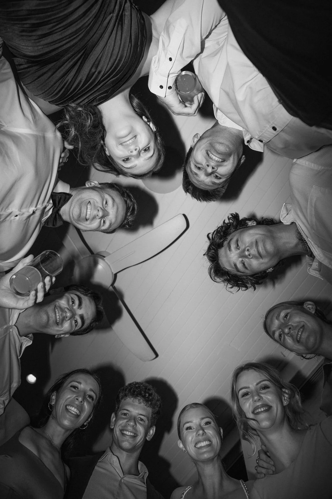 Group of friends smiling and looking down at the camera in a circle at a party or gathering, with a ceiling fan overhead.
