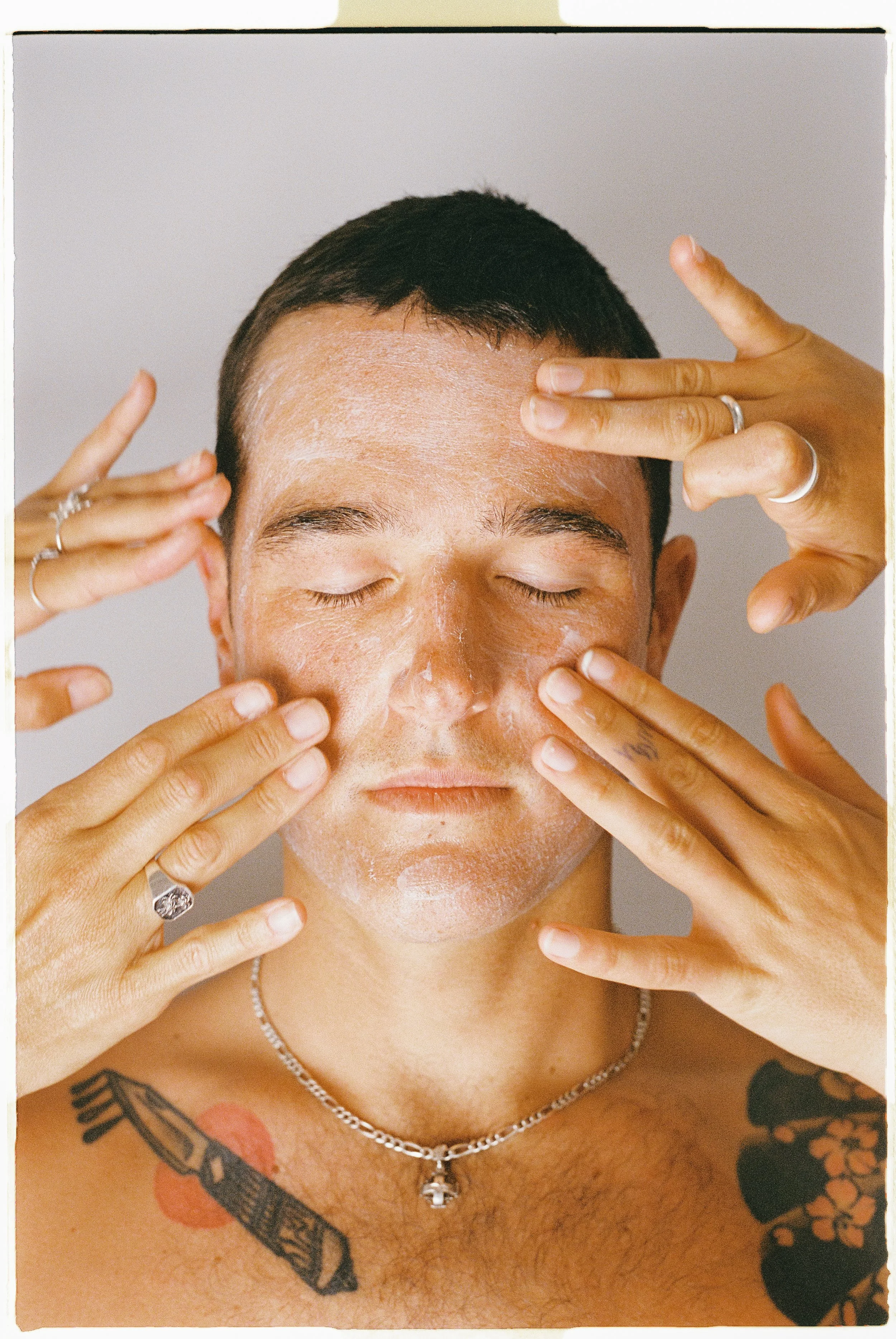 Person with short dark hair, closed eyes, applying or massaging a white substance, possibly cream or face mask, on face with multiple hands.