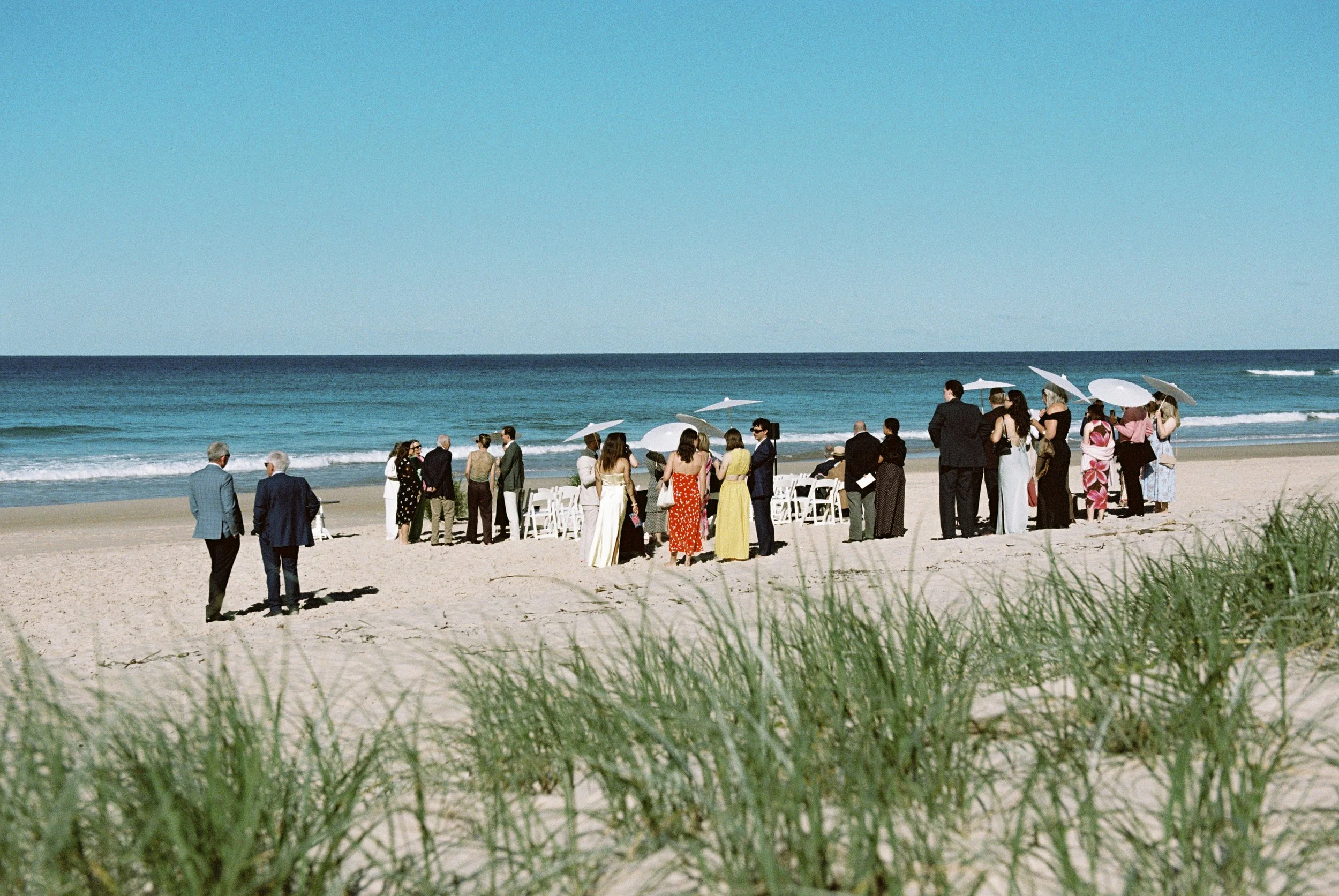 A group of people gathered on a beach for a wedding ceremony with white chairs, umbrellas, and a view of the ocean under a clear blue sky.