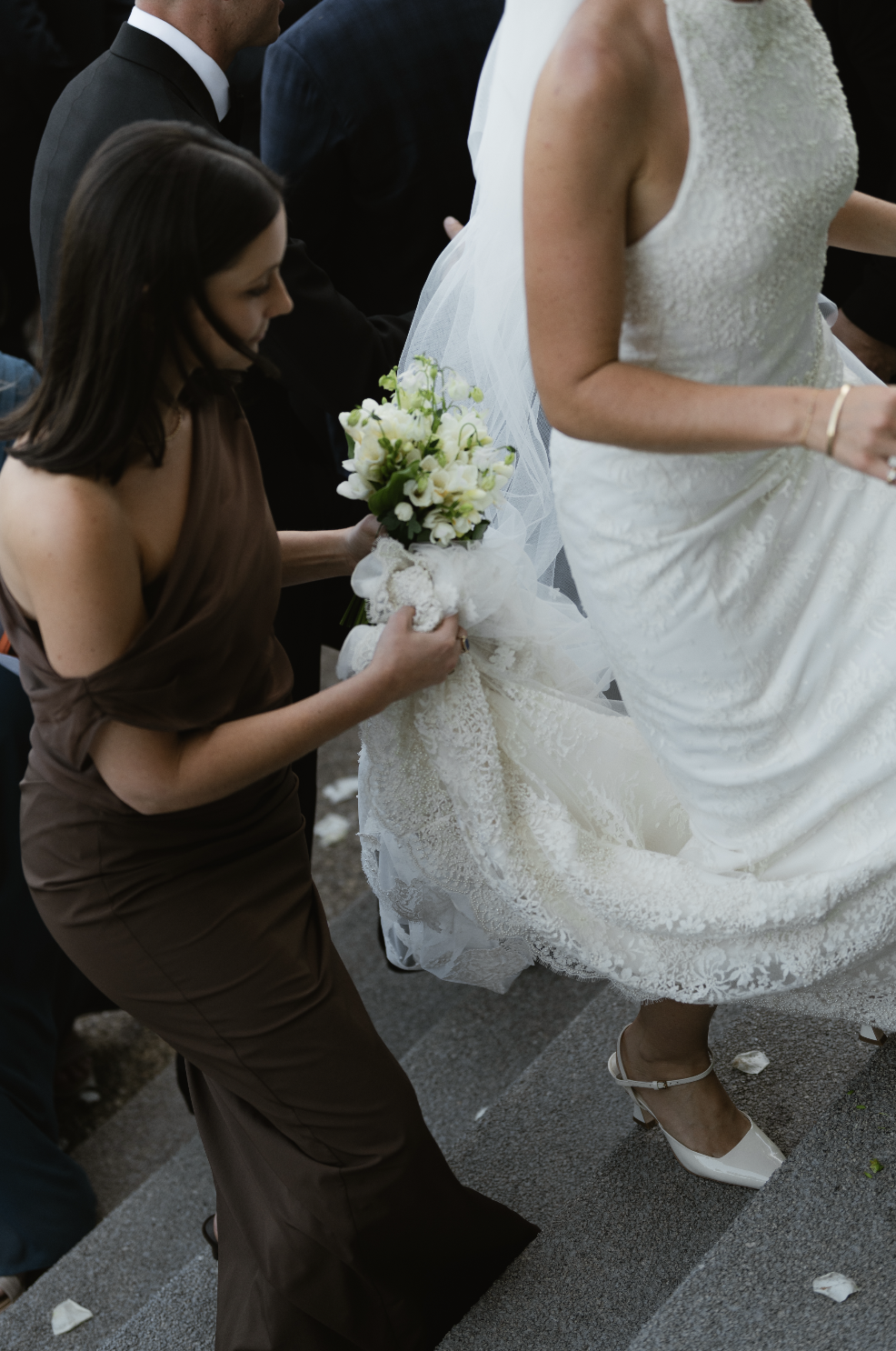 A bride in a white wedding dress is being helped by a woman in a brown dress to hold her gown and bouquet during a wedding ceremony.