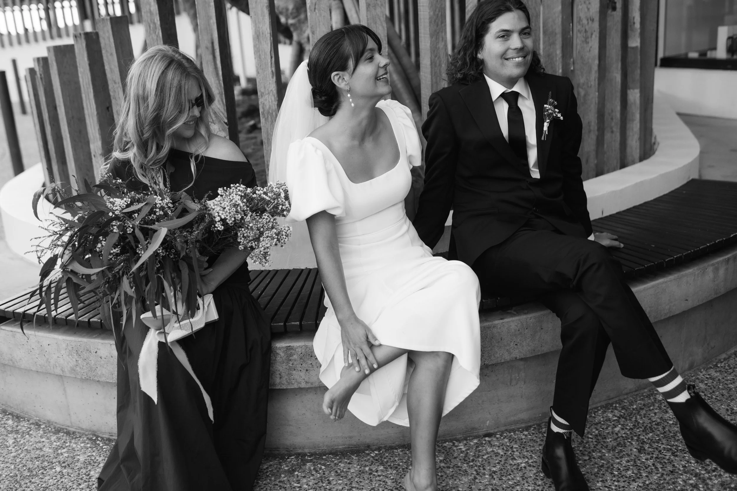 Black and white photo of three people sitting on a curved bench. Two women and a man wearing formal attire. The woman in the middle is in a white dress, smiling, with her hand intertwined with a person outside the frame. The woman on the left wears g