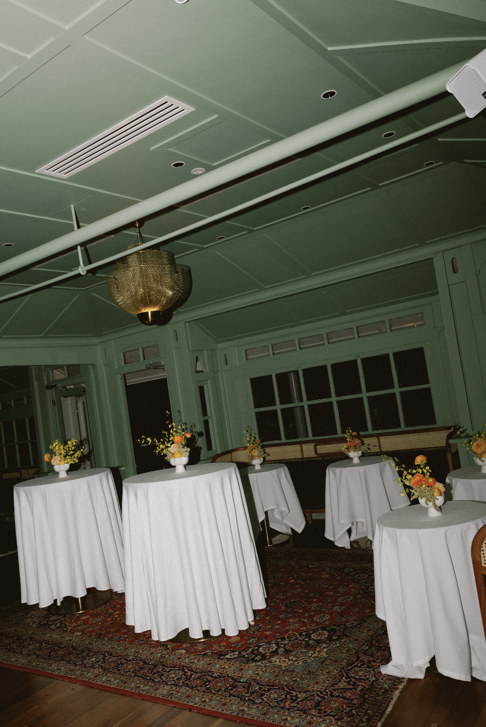 An elegant event space with green paneled walls, high ceiling, and large windows. Several round tables covered with white tablecloths are decorated with floral centerpieces. A vintage-style carpet is on the wooden floor, and a gold chandelier hangs f