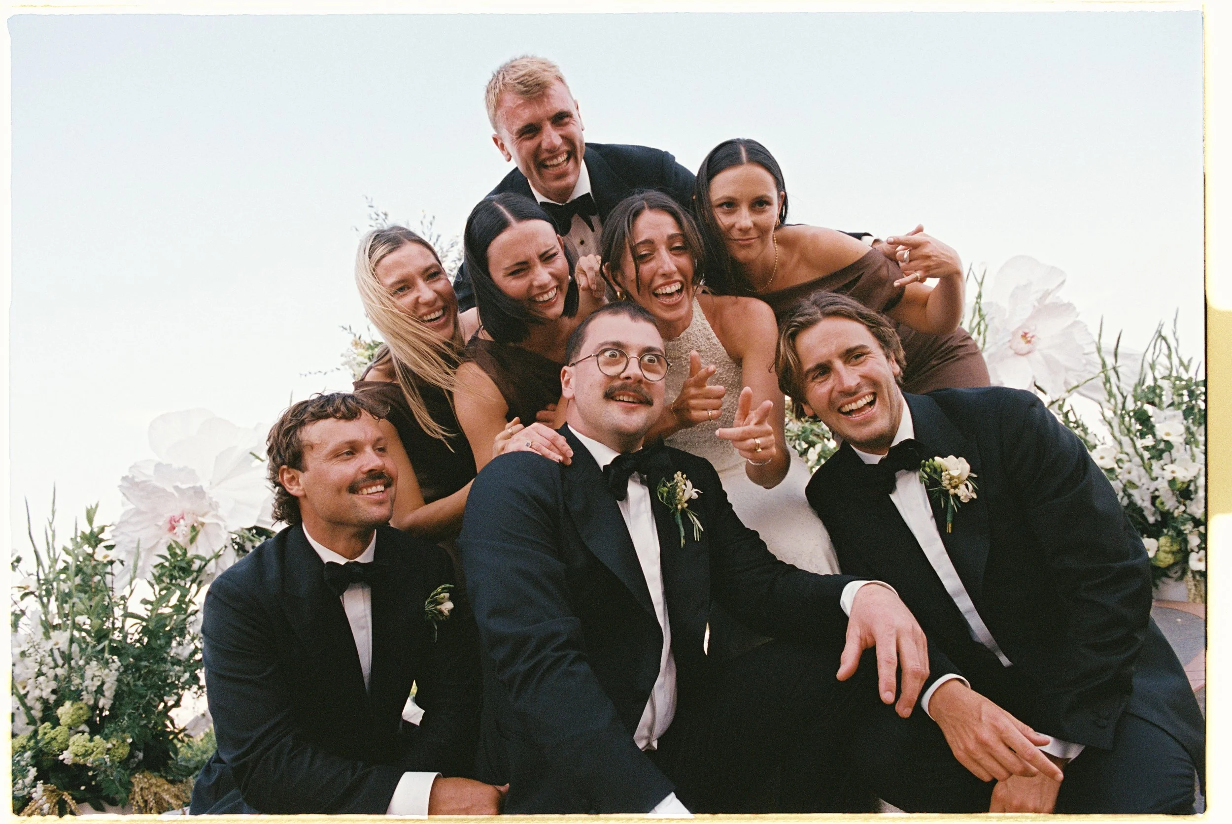 Group of friends in formal wedding attire, smiling and making playful gestures, posed outdoors against a floral backdrop.