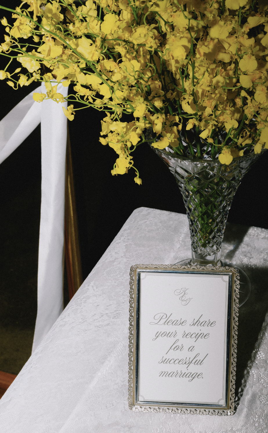 A tall crystal vase holding yellow flowers on a white table with a framed sign that reads, 'Please share your recipe for a successful marriage,' at a wedding or special event.