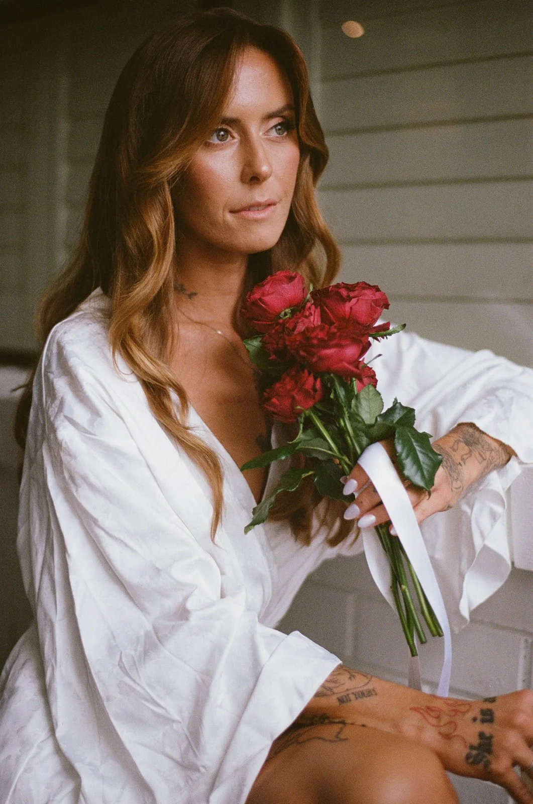 A woman with wavy brown hair and tattoos on her arms, wearing a white robe, holding a bouquet of red roses, sitting near a wall, looking thoughtfully.