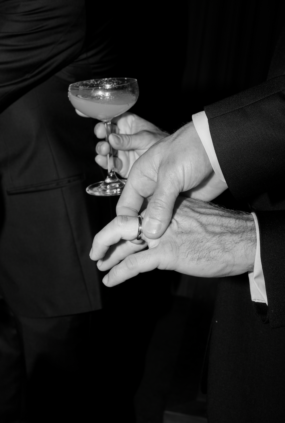 Close-up of a couple holding hands, with one person wearing a wedding band, at a formal event. The person in the background is holding a coupe glass with a beverage.