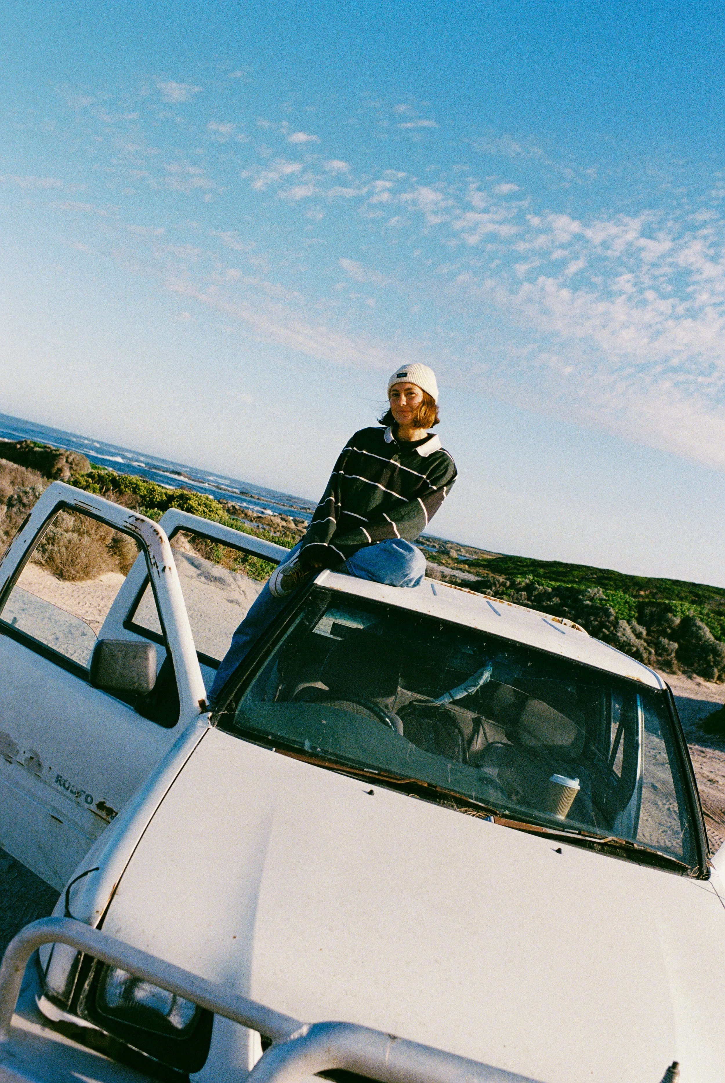 A woman with brown hair wearing a white knit beanie, black and white striped sweater, and jeans sitting on top of a white Jeep near a coastline with rocks, bushes, and the ocean in the background under a cloudy blue sky.