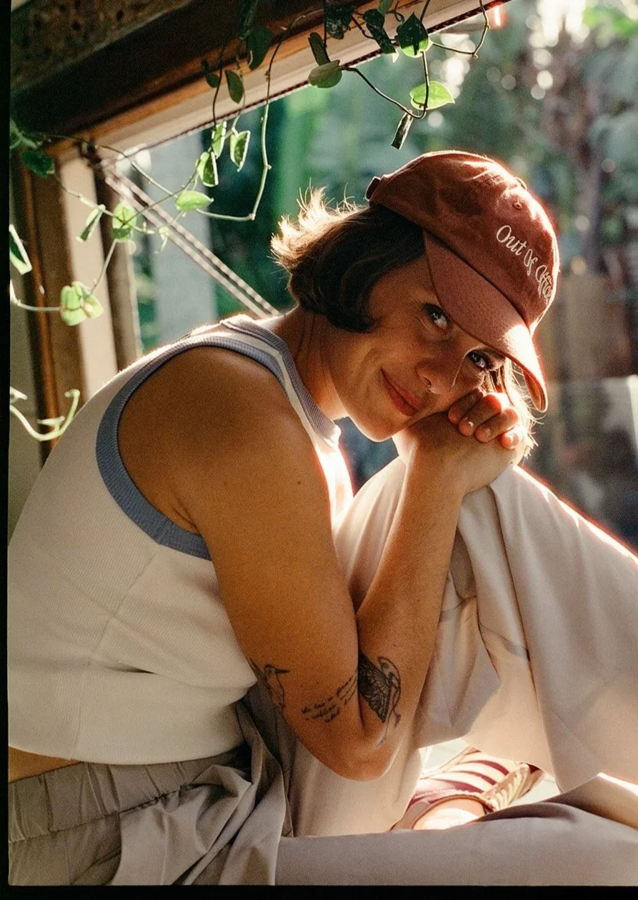 A woman sitting and smiling, wearing a red cap and sleeveless white shirt with blue trim, with tattoos on her arm, outside during sunlight, with greenery and string lights in the background.