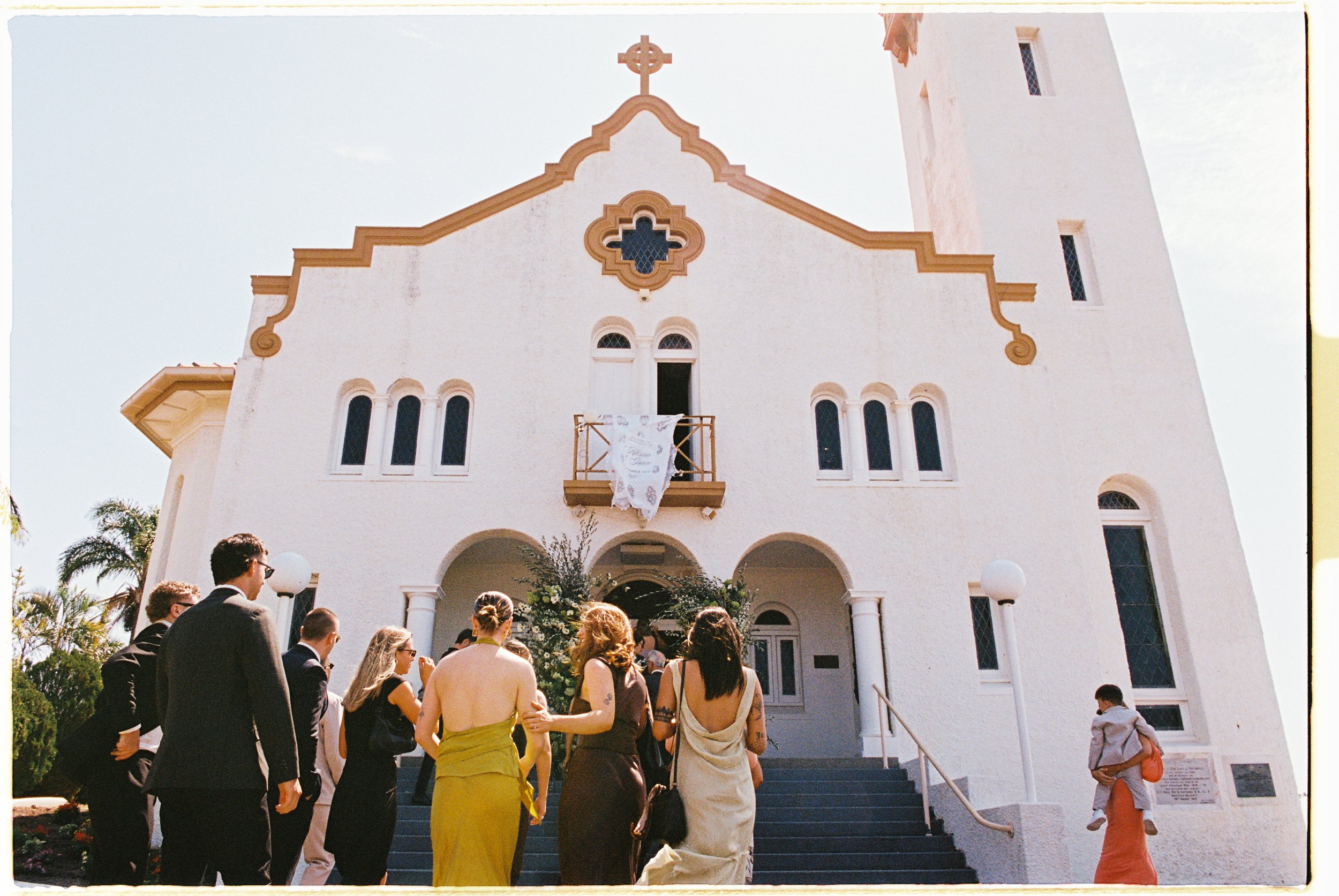 People attending a wedding ceremony outside a white church with a steeple and cross on top, decorated with flowers and white fabric.