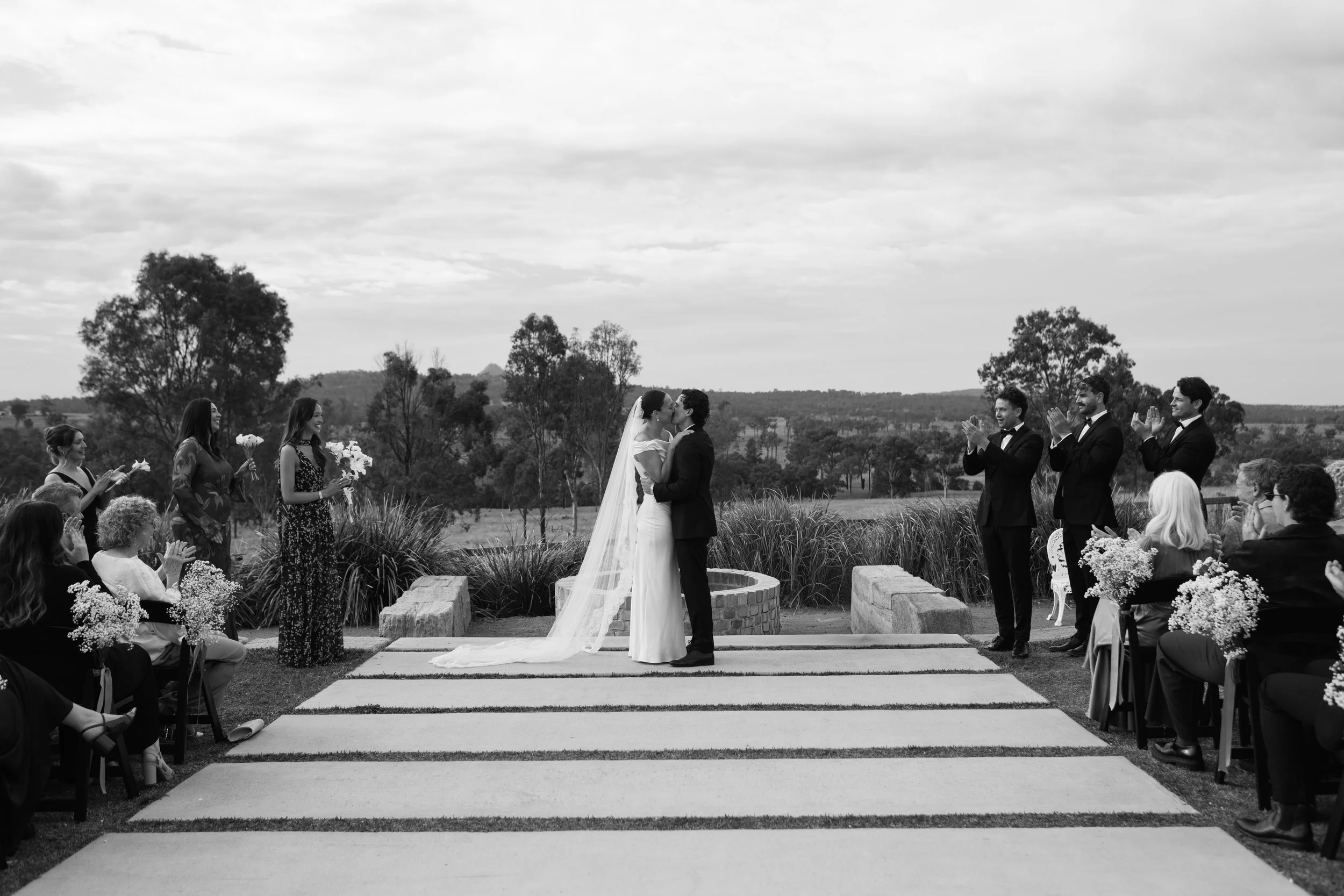 A black and white photo of a outdoor wedding ceremony with the bride and groom kissing, surrounded by guests seated on both sides, with a scenic landscape in the background.