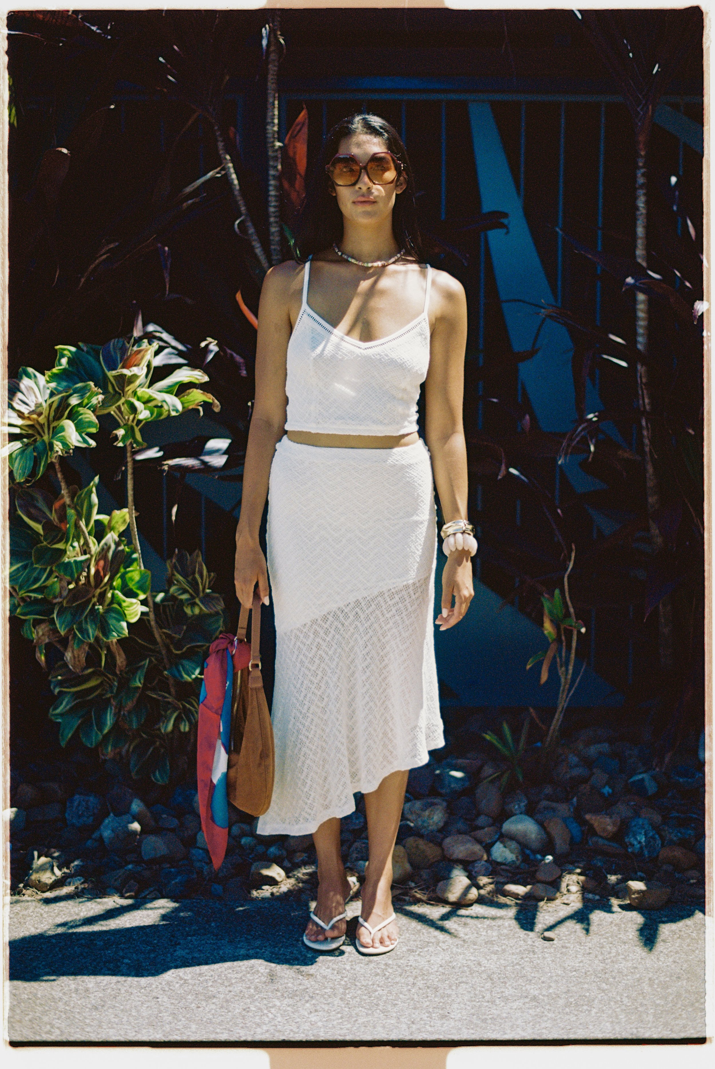 A woman wearing a white crochet crop top and skirt standing outdoors in sunlight, holding a bag with a red and pink scarf, wearing sunglasses, jewelry, and sandals, with tropical plants and a blue fence in the background.