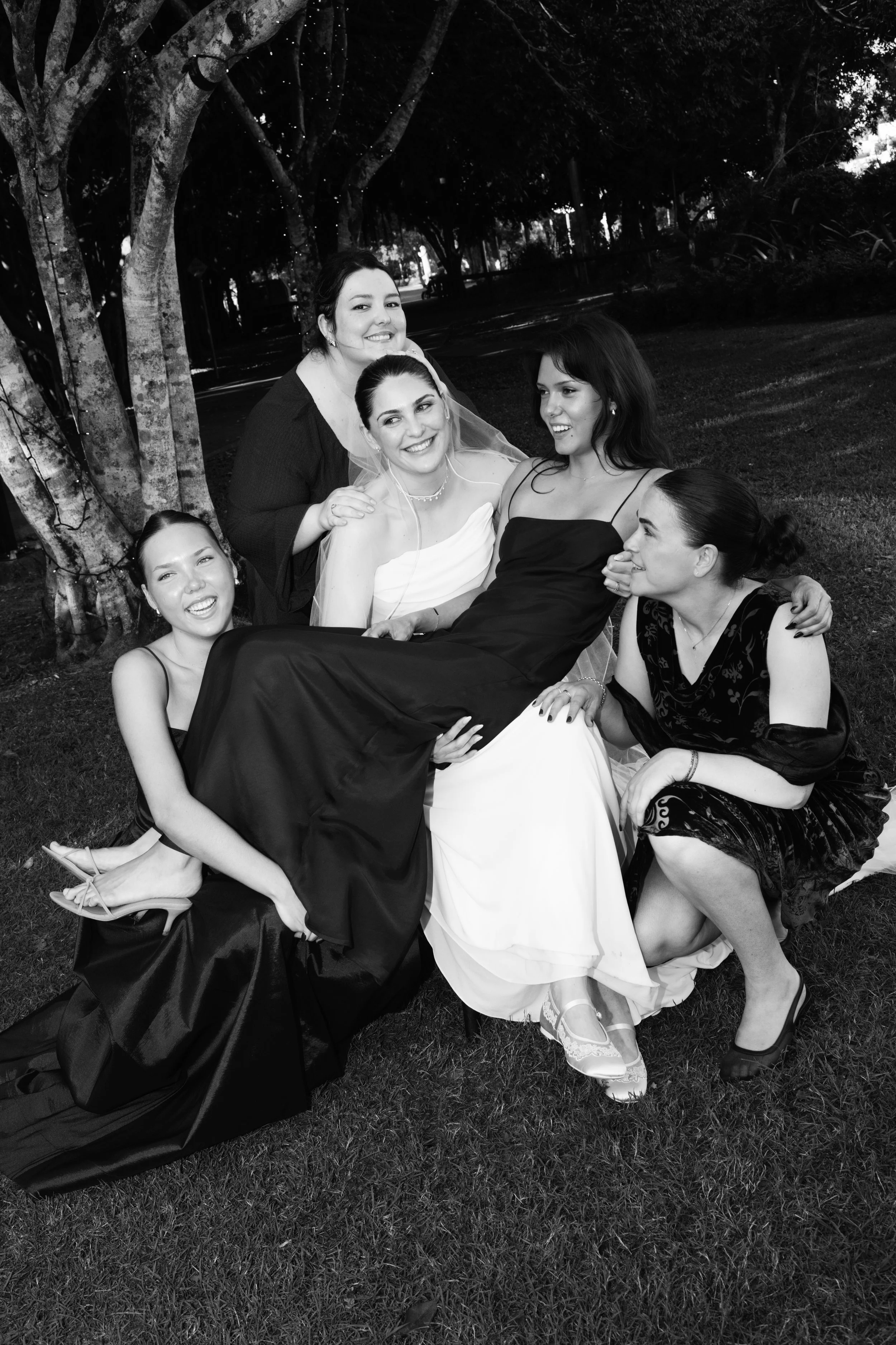 Black and white photo of five women sitting outdoors under trees, with one woman in a wedding dress, others in formal attire, smiling and interacting with each other.