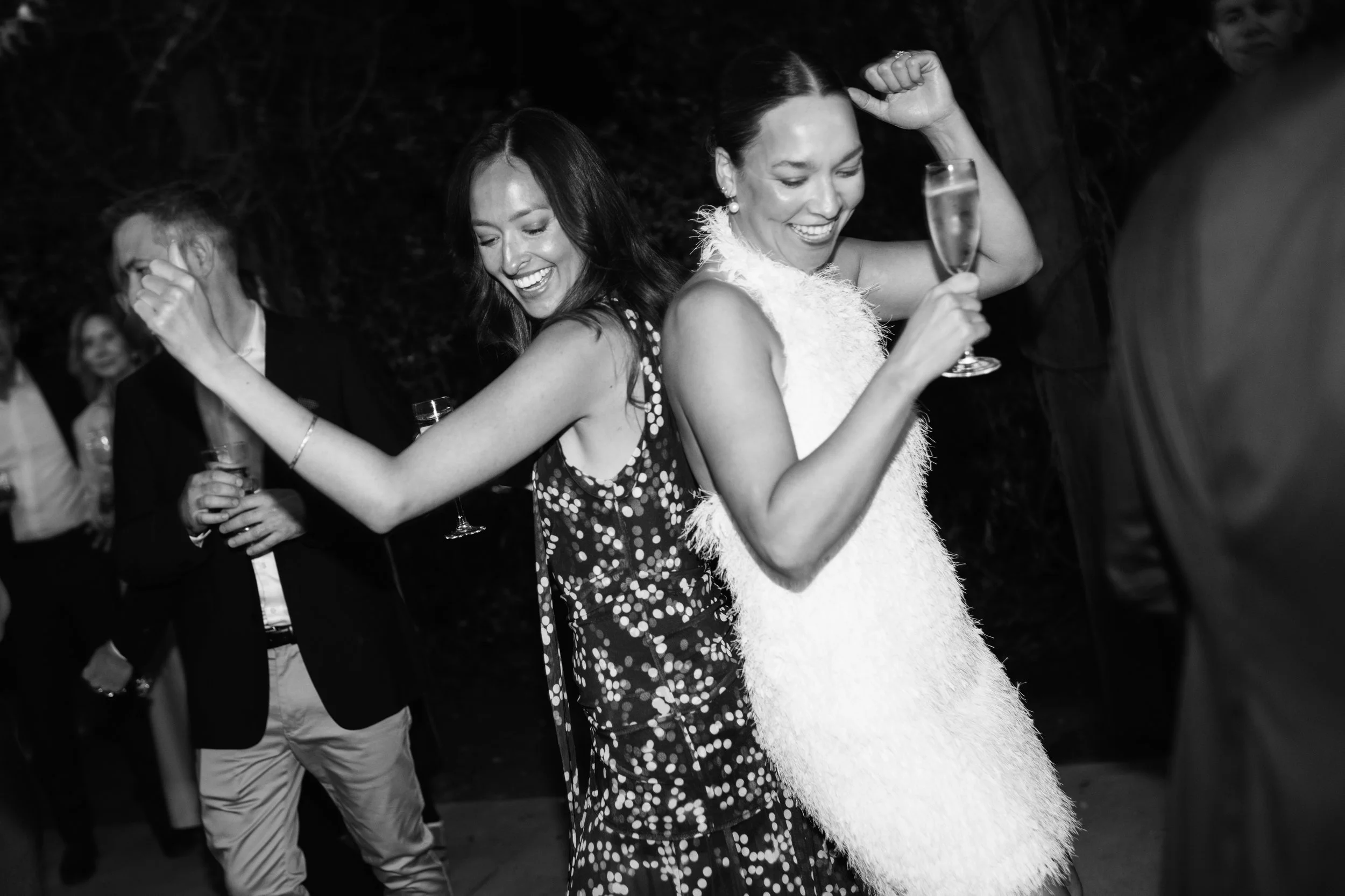 Friends dancing and enjoying themselves at a celebration, with women smiling and holding drinks, in black and white.
