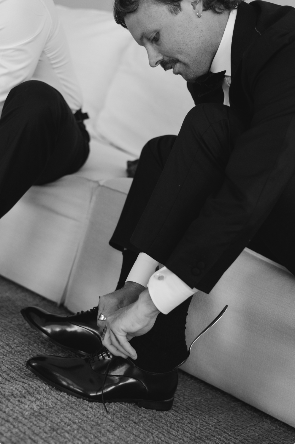Man in tuxedo putting on black dress shoes while sitting on a sofa.