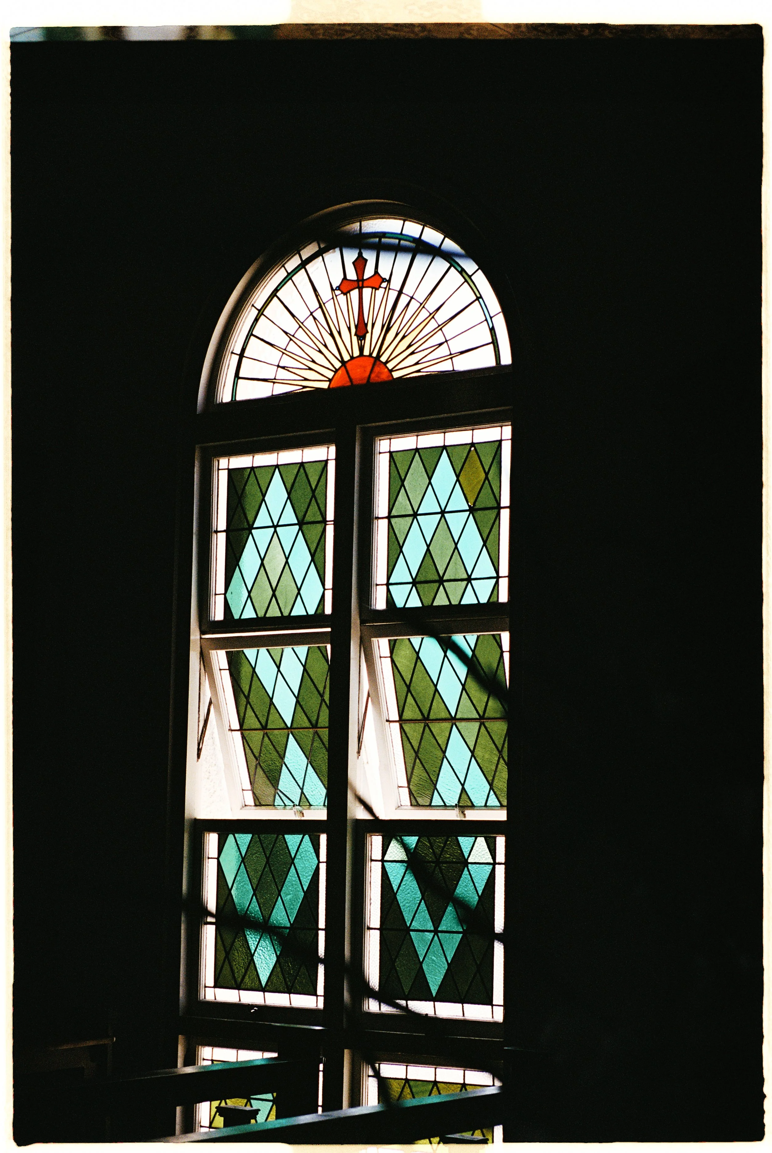 Stained glass window with geometric green and blue patterns and a semicircular top with orange accents and clock hands.