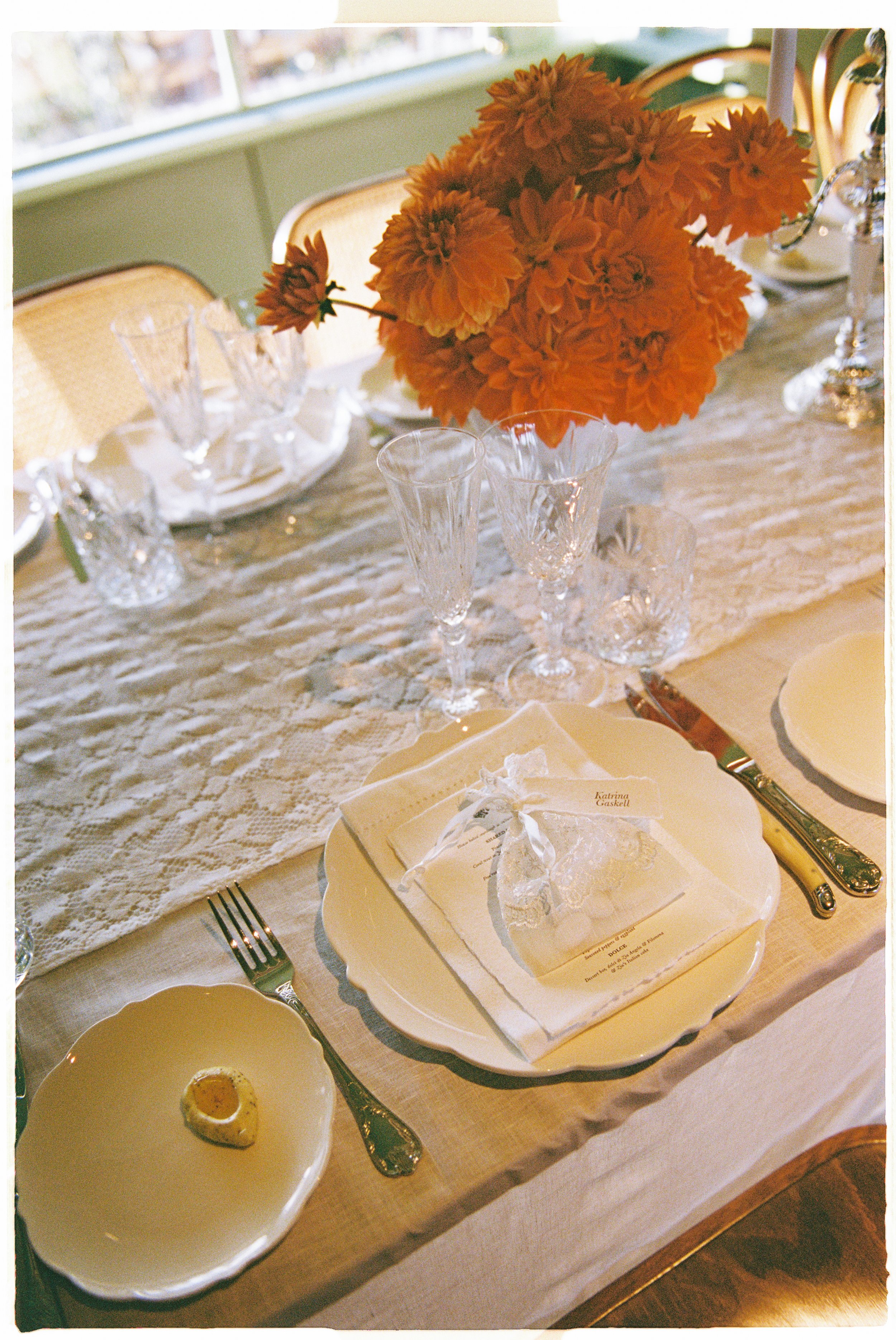 A dining table set with a white tablecloth, a large bouquet of orange flowers, crystal glassware, and a place setting with a white plate, a fork, and a napkin with a wedding favor ribbon. There is a small bowl with an egg and sauce. In the background, there are empty yellow chairs and a window.