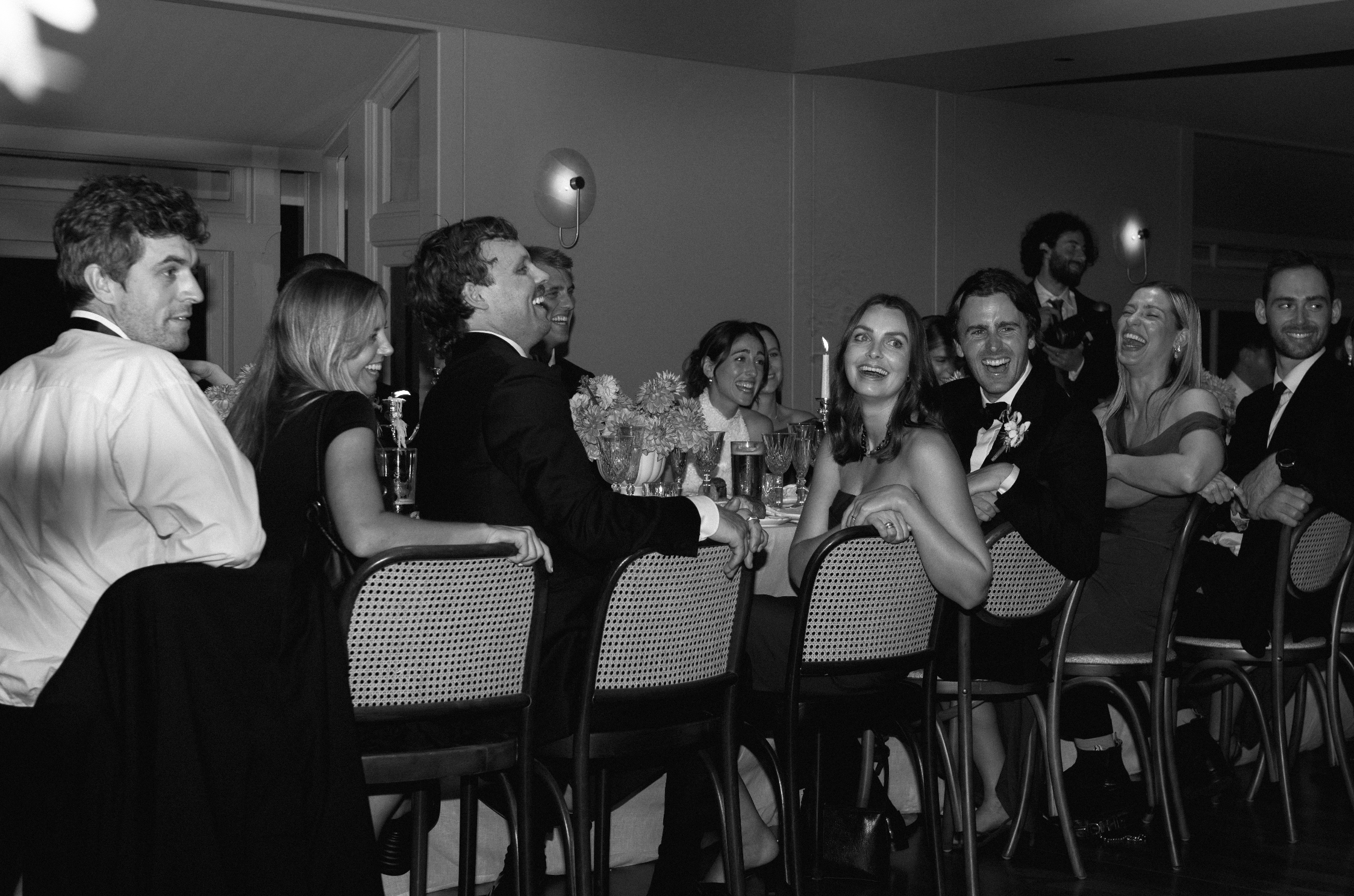 A group of people dressed formally sitting and standing around a long table at a celebration, all smiling and laughing.