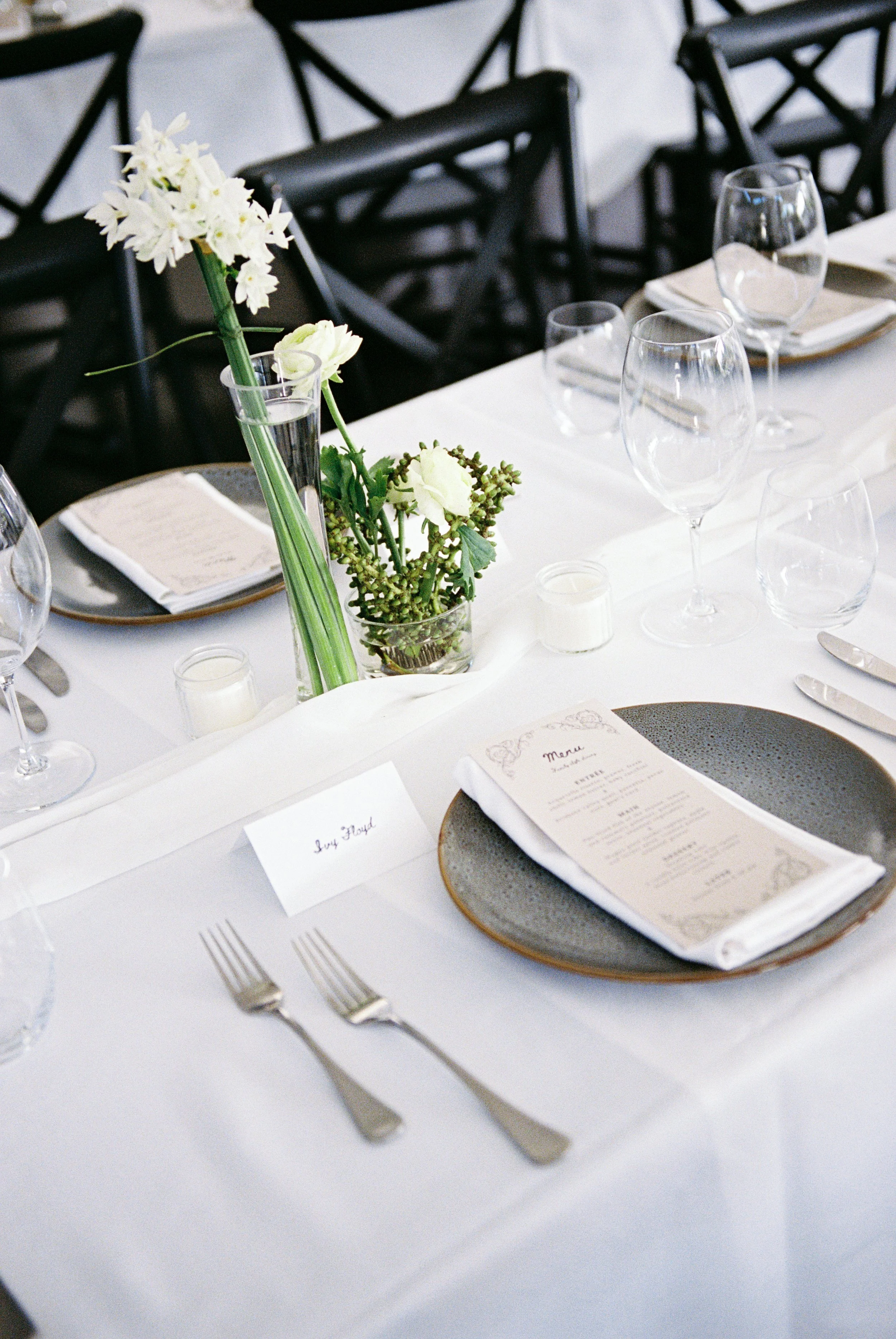 A formal dining table set with white tablecloth, wine glasses, silverware, a menu, and a centerpiece of white flowers in a tall vase, with black chairs in the background.