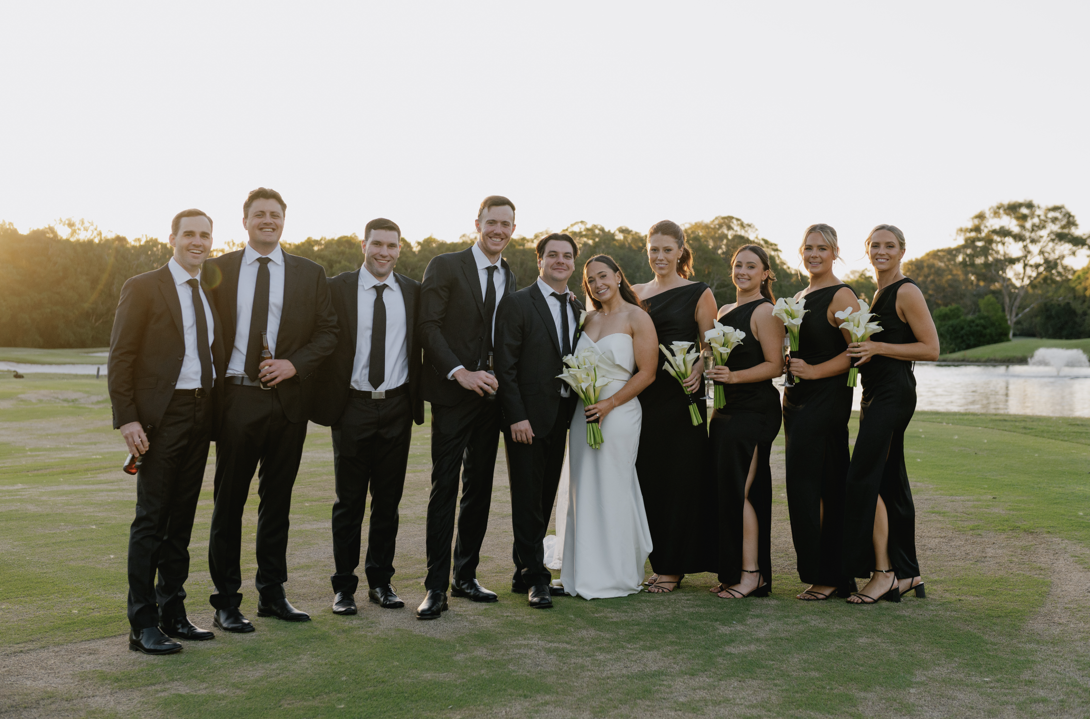 Group of wedding guests and bride posing outdoors near a pond at sunset, holding flowers and drinks.