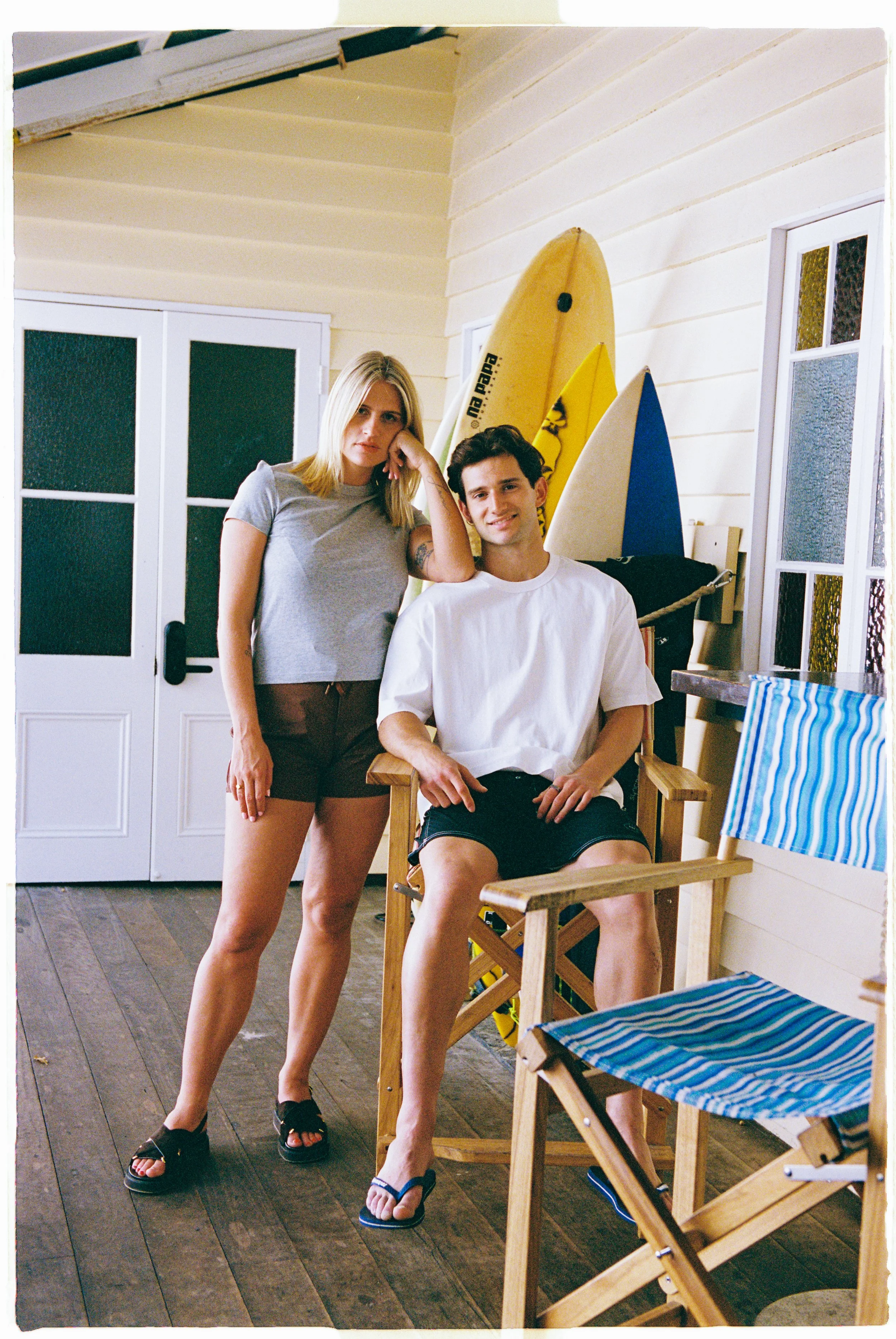 A woman standing next to a seated man inside a beach house, with surfboards behind them and a wooden chair and striped beach chair nearby.