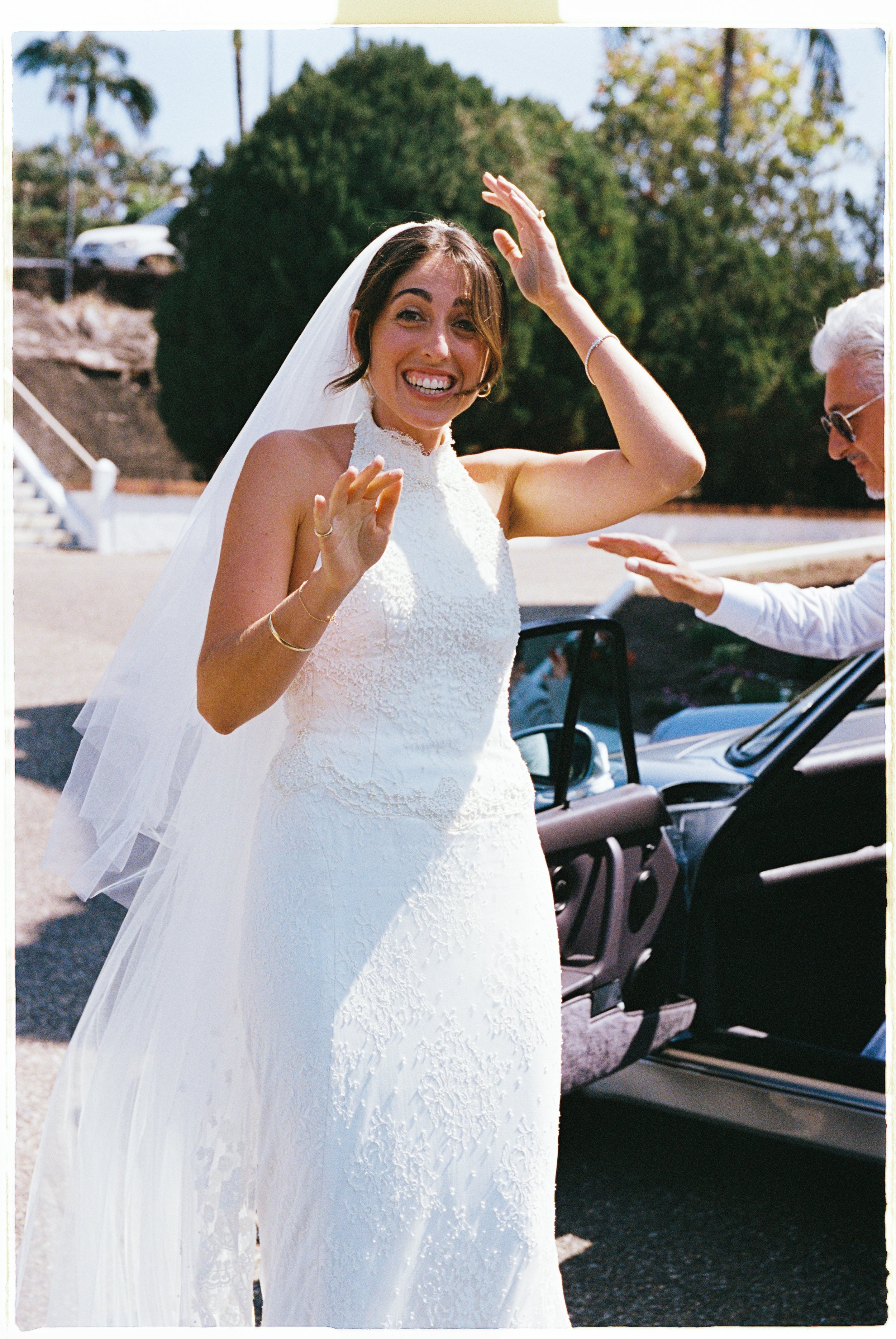 A smiling bride in a white lace wedding dress and veil standing outside near a car, waving or about to wave, with a person reaching out to her in the background.