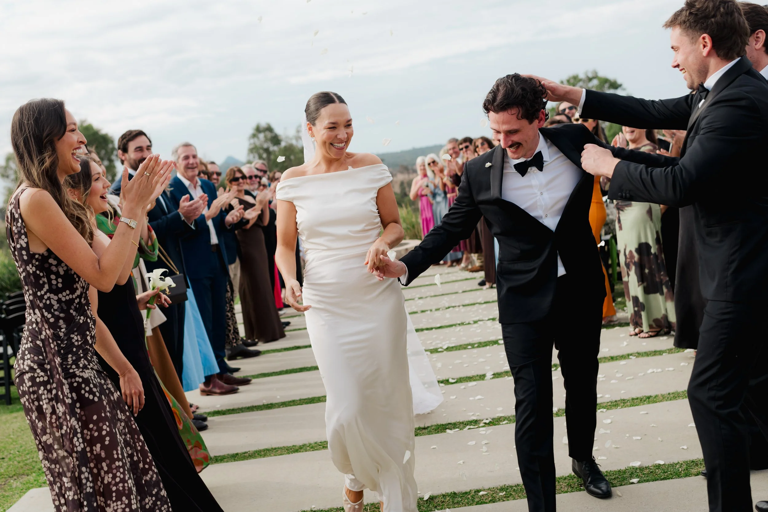 A group of people celebrating a wedding outdoors, with the bride and groom walking happily, surrounded by friends clapping and smiling, some throwing flower petals.