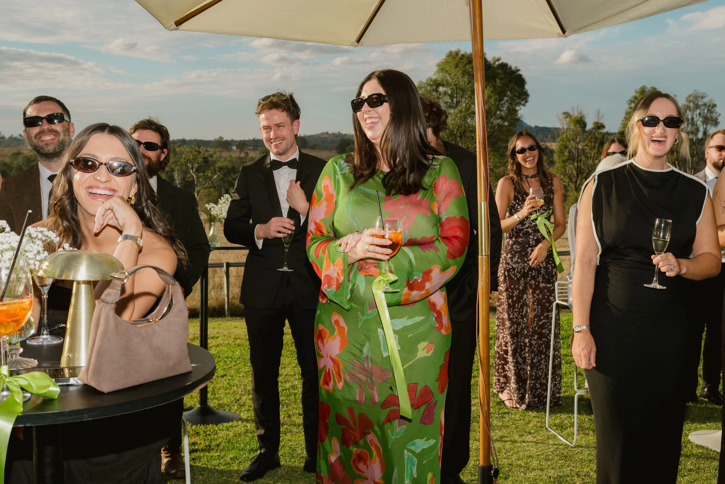 Group of people at an outdoor celebration, some holding drinks, under a large umbrella on a sunny day with scenic landscape in background.