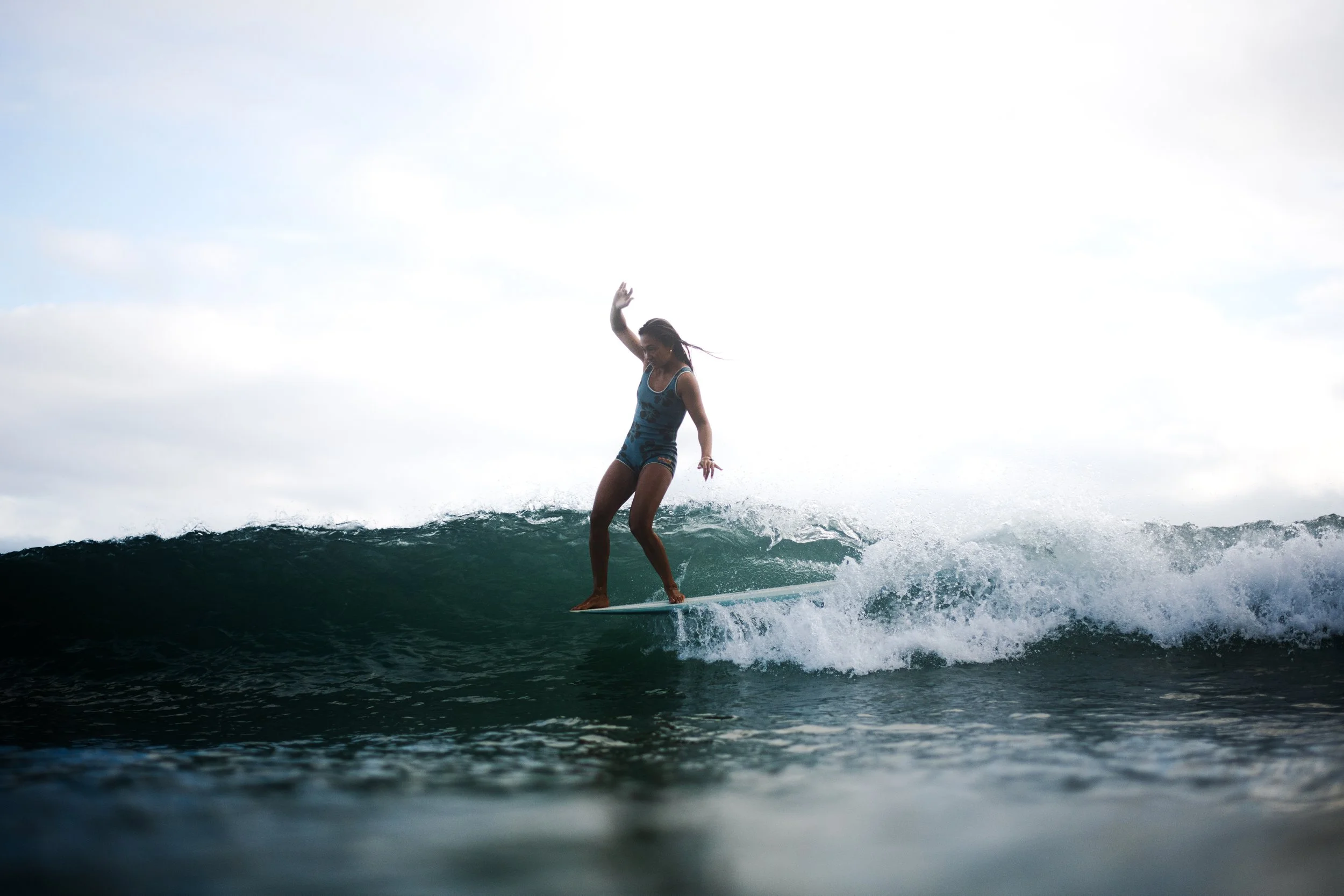 Woman surfing on a wave in the ocean, wearing a blue swimsuit, with a cloudy sky in the background.