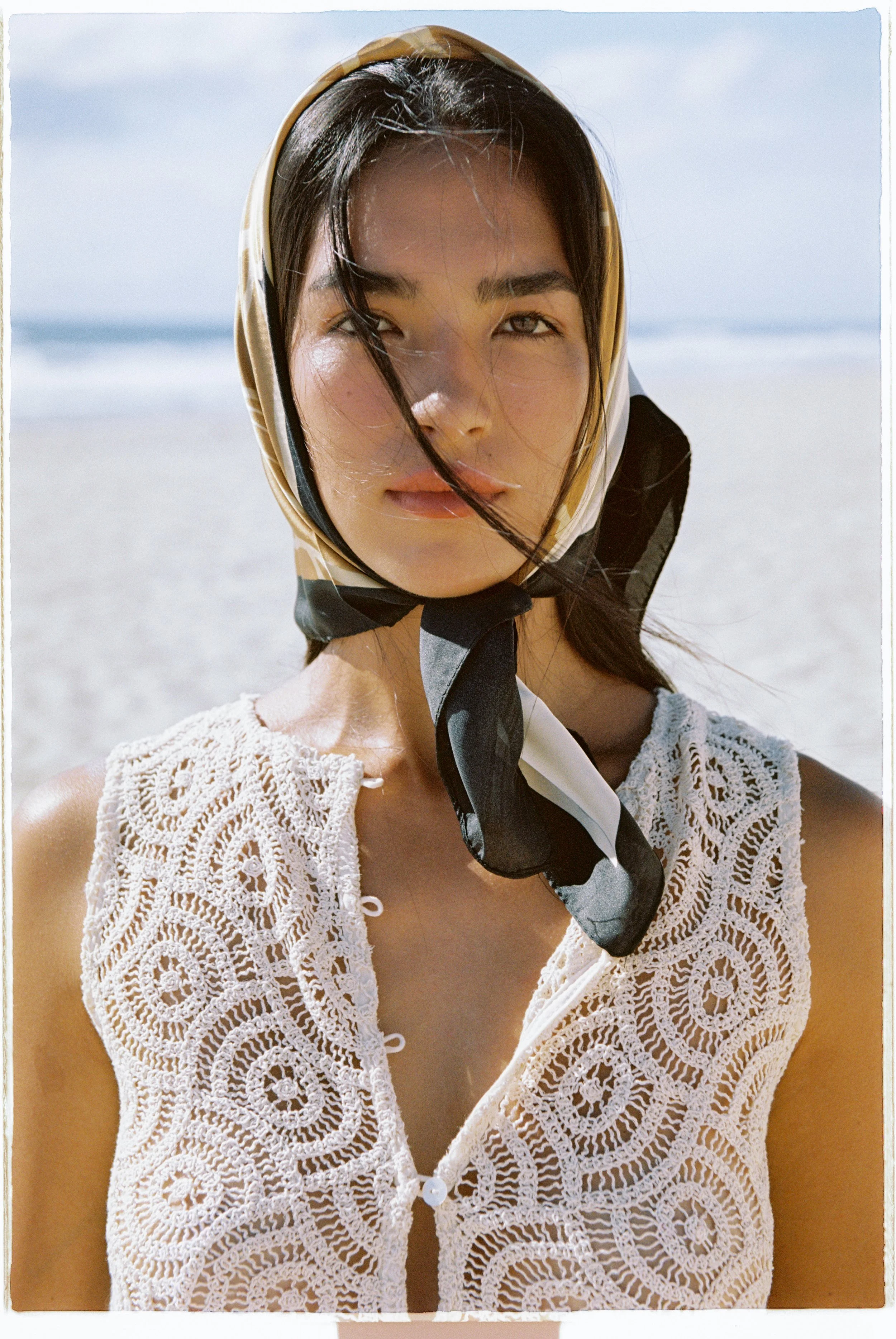 A young woman with black hair wearing a patterned headscarf, a sleeveless crochet top, and sitting on a beach with ocean waves in the background.