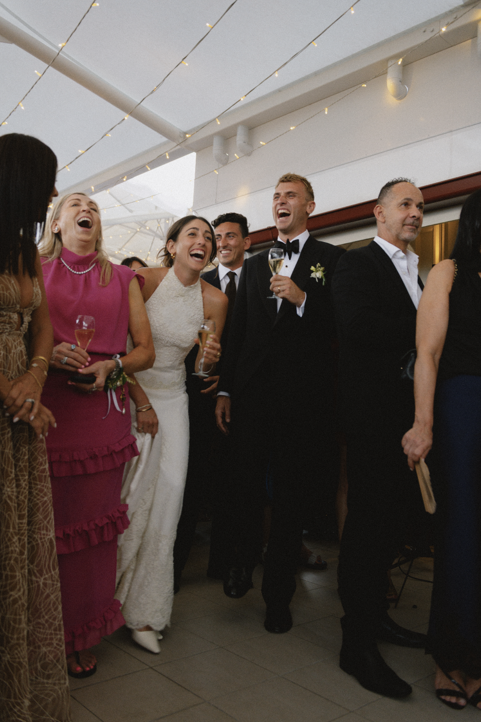 Group of people at a wedding reception, smiling and laughing, dressed in formal attire with some holding champagne glasses.