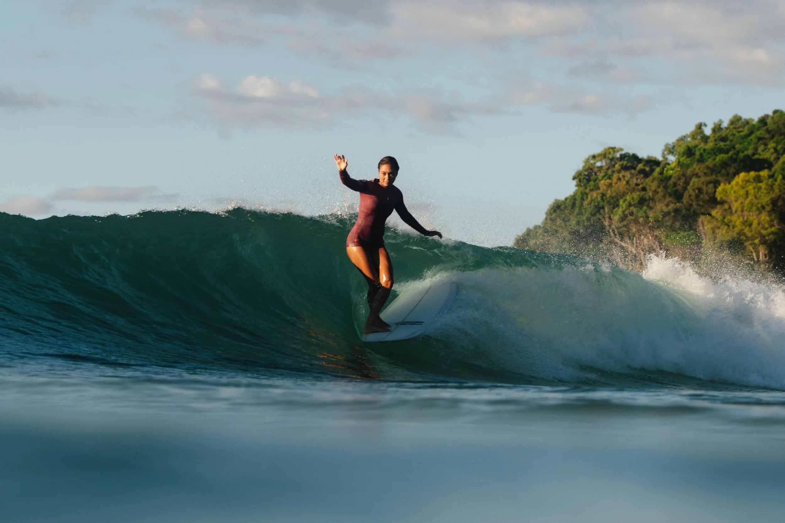 Lilliana Bowrey surfing on a wave in the ocean near a green, tree-covered island under a partly cloudy sky, wearing Billabong