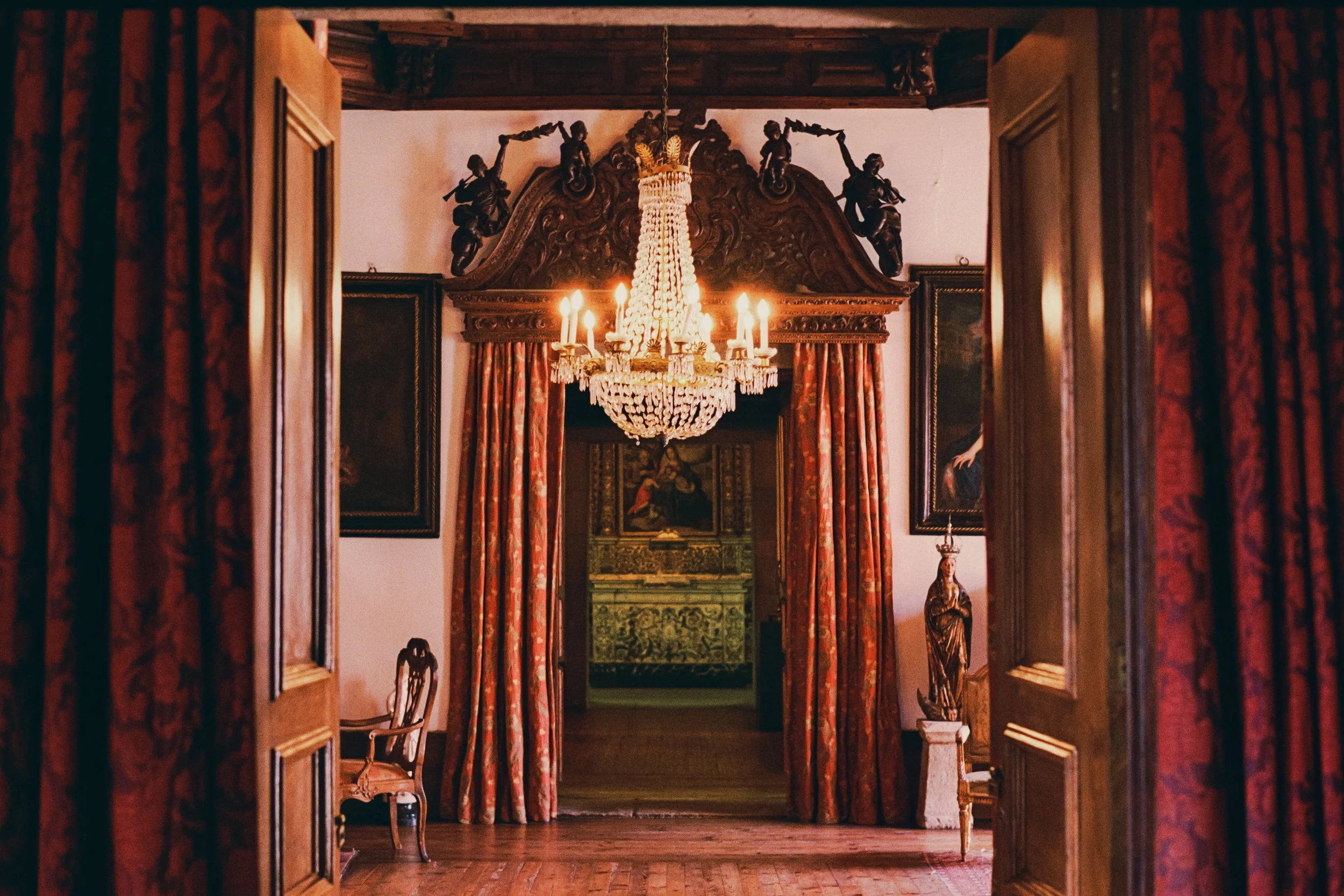 An elegant chandelier hangs from the ceiling of a richly decorated room, viewed through open wooden doors with carved detailing. The room has red and gold curtains, ornate wooden furniture, and a framed painting.