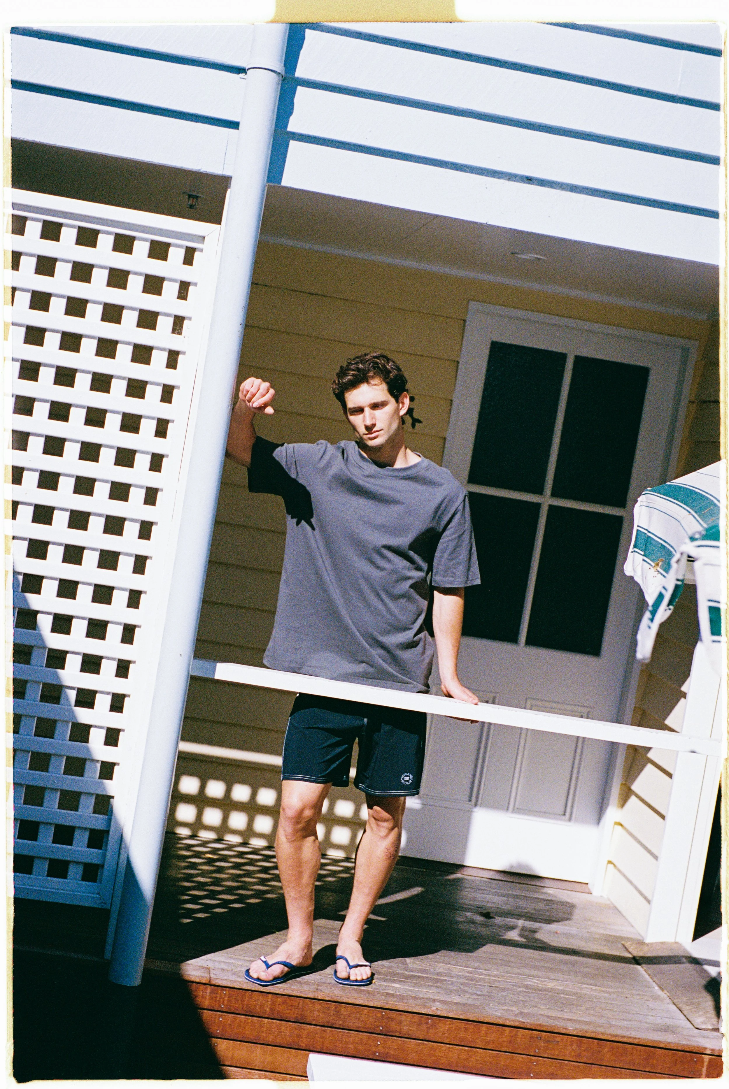 A young man standing on a wooden porch, wearing a gray T-shirt, navy shorts, and flip-flops, leaning against a white railing with a house in the background.