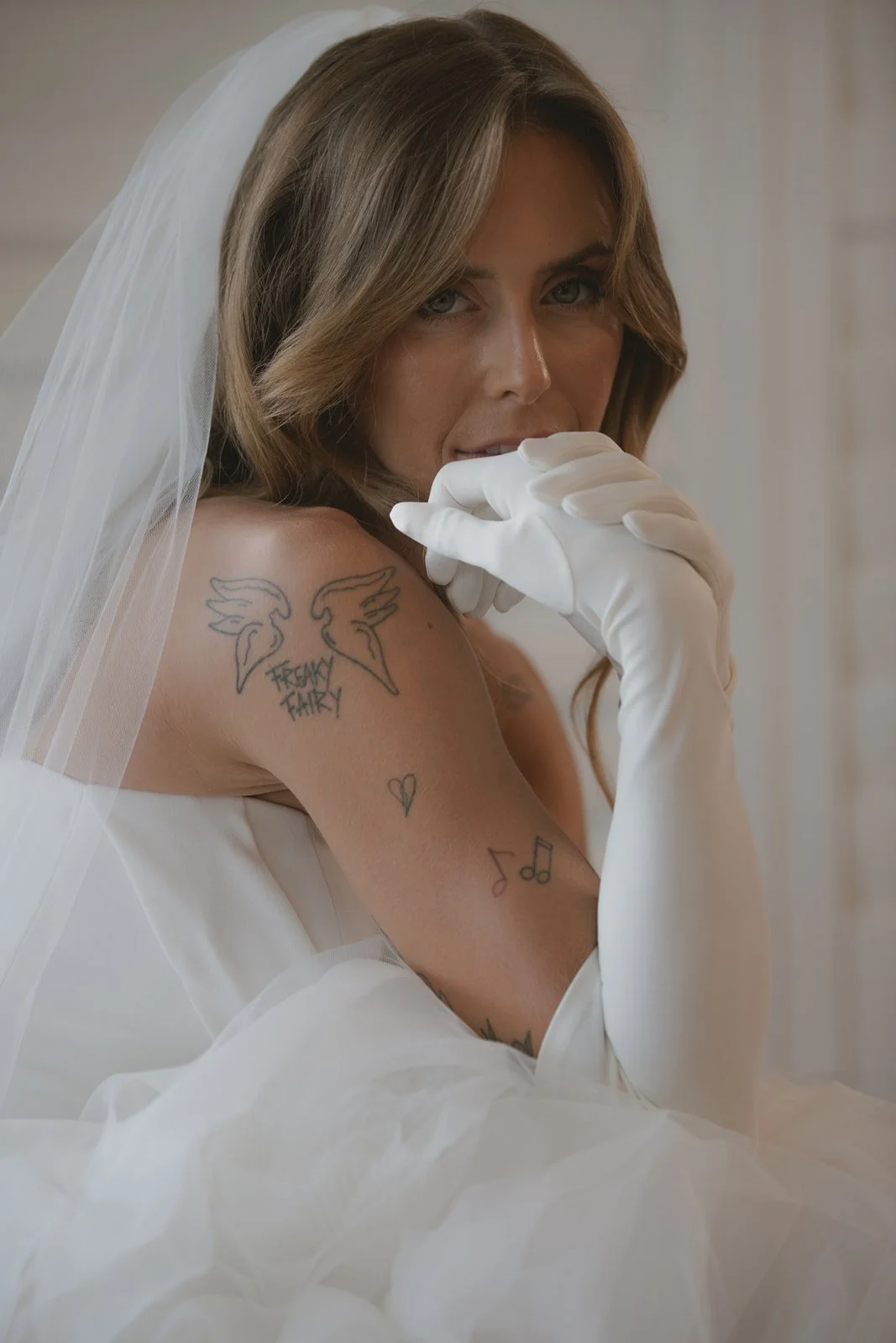 A woman with tattooed arm wearing a bridal veil and white gloves, looking at the camera.