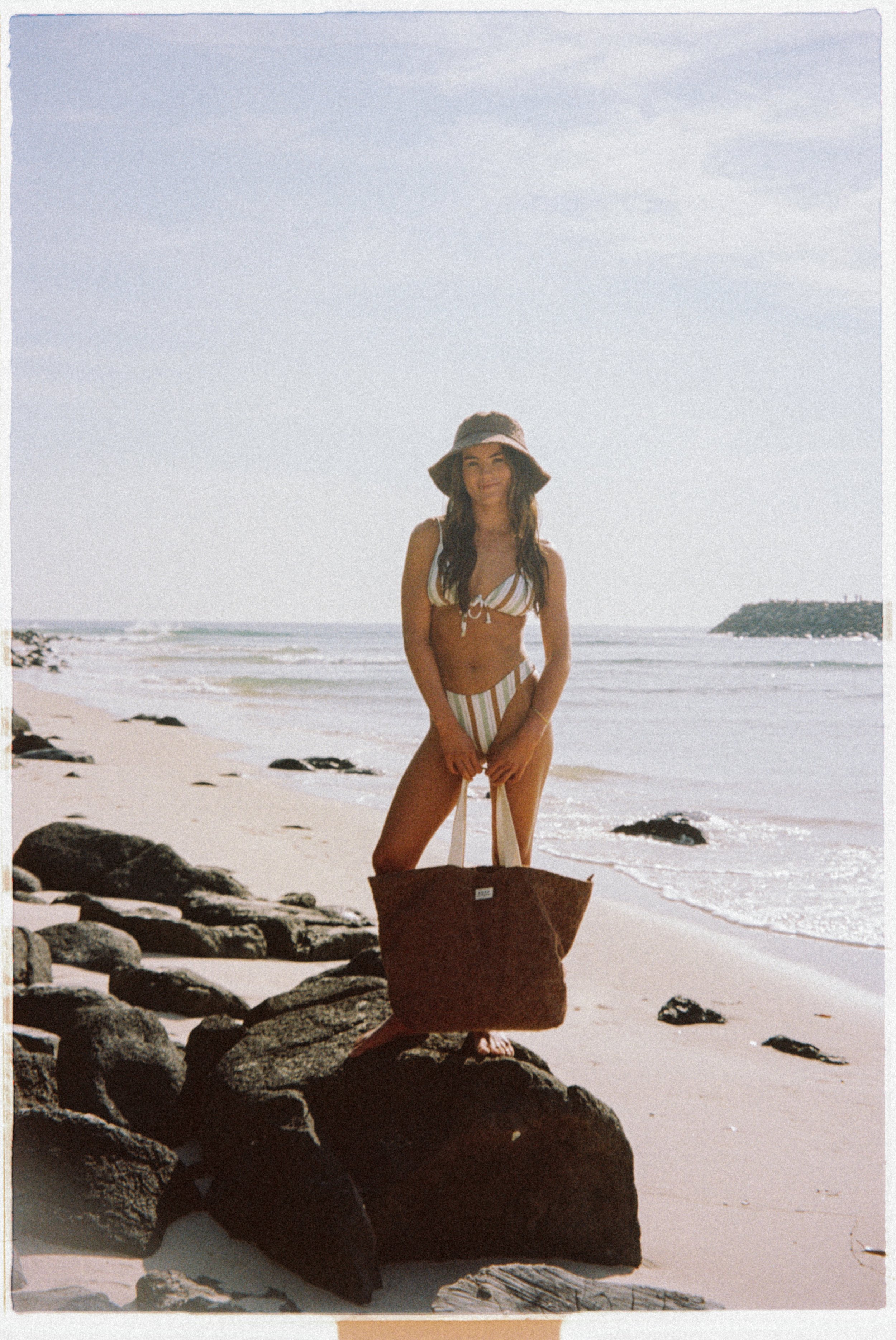 A woman in a striped bikini and sun hat stands on rocks on a beach with the ocean and sky in the background, holding a large tote bag.
