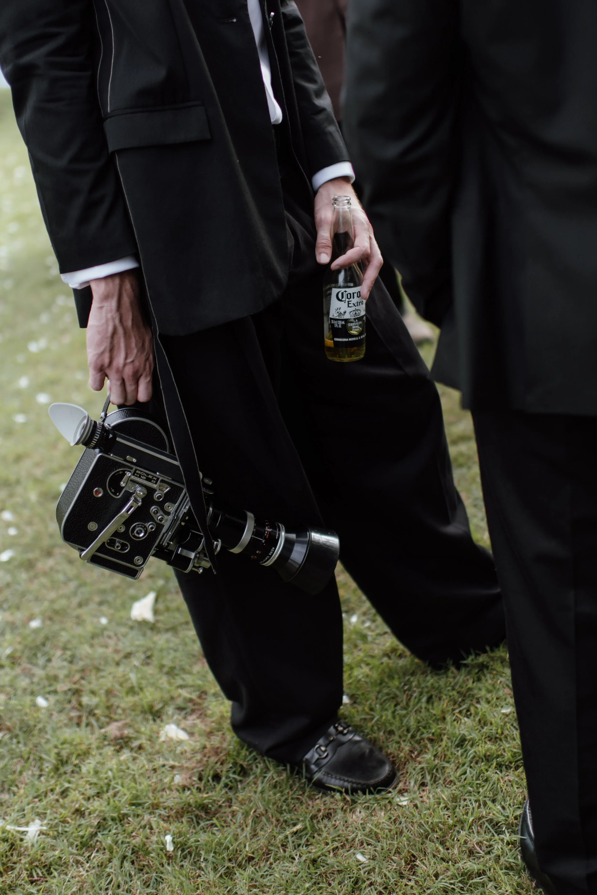 Close-up of a person wearing a black suit holding a bottle of Corona beer in one hand and a vintage film camera in the other, standing on grass with scattered white petals.