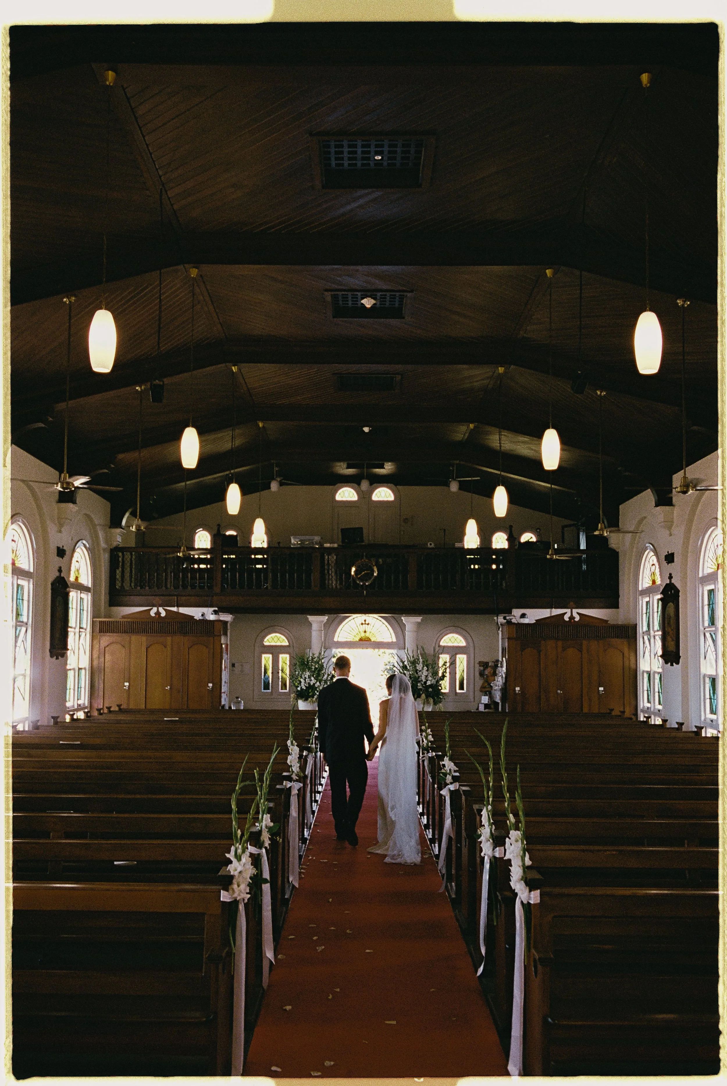 A bride and groom walking down the aisle of a church decorated for a wedding, with white ribbons and flowers on the pews, leading toward the altar at the front.