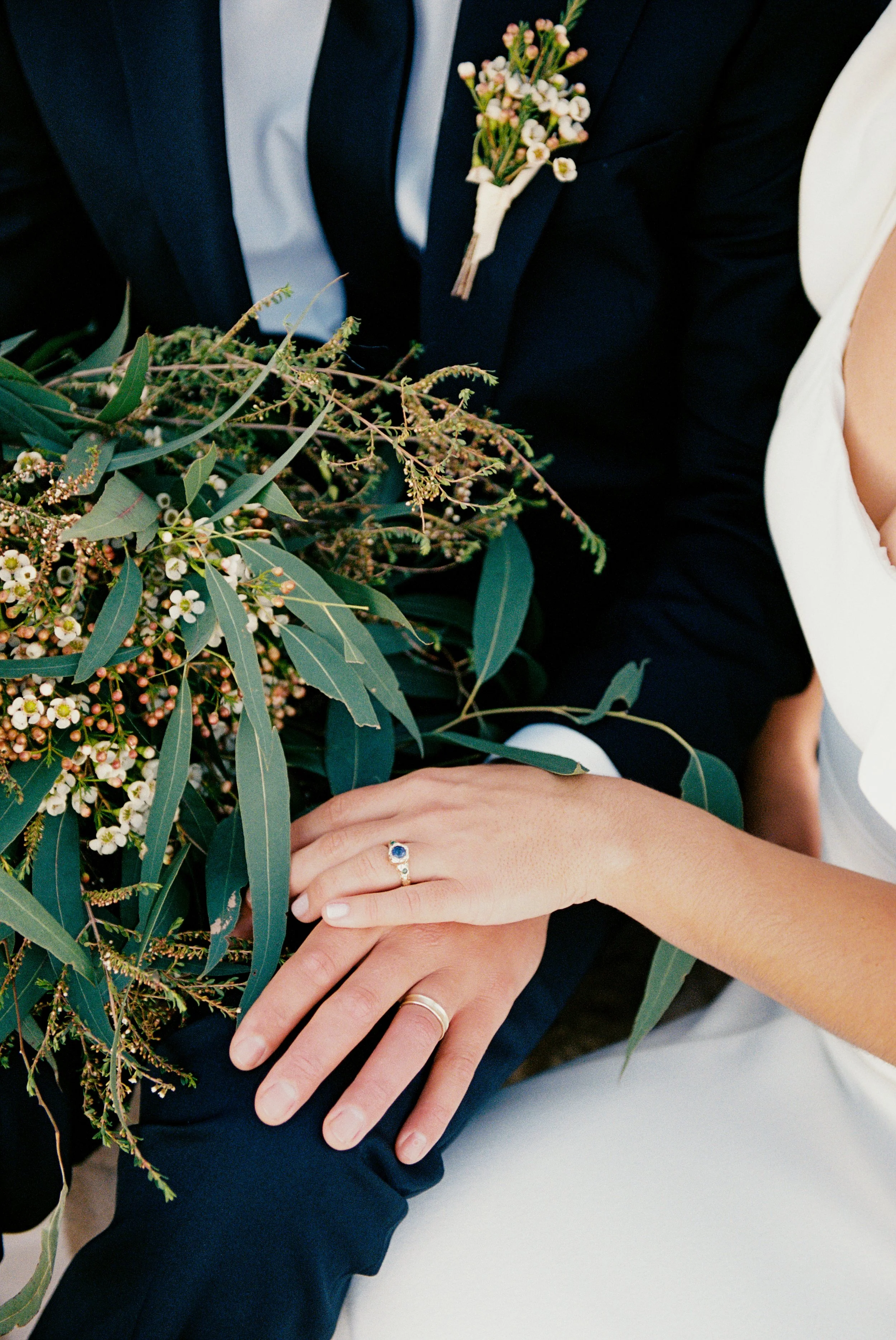 Close-up of a bride and groom's hands, showing wedding rings, with a bouquet of greenery and small white flowers, during a wedding ceremony.