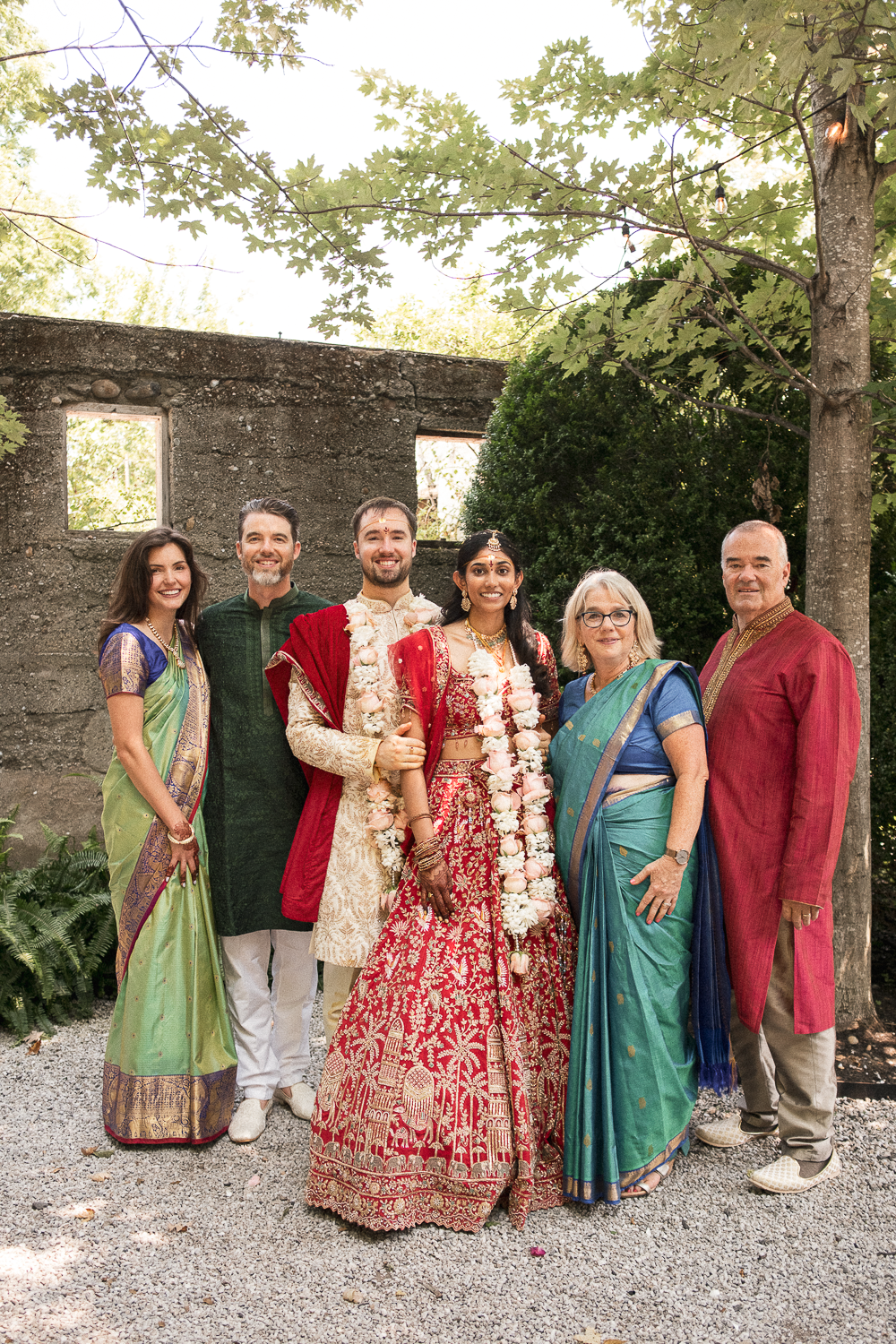 A wedding party standing outdoors with six people, including the bride and groom dressed in traditional Indian wedding attire, surrounded by family members in colorful sarees and kurta. The background features a stone wall and lush green trees.