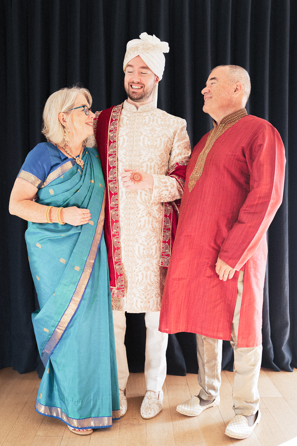 Three people dressed in traditional Indian attire smiling and conversing in front of a black curtain. The woman on the left wears a turquoise saree with gold accents and glasses, the man in the middle wears a cream and red sherwani with a turban, and