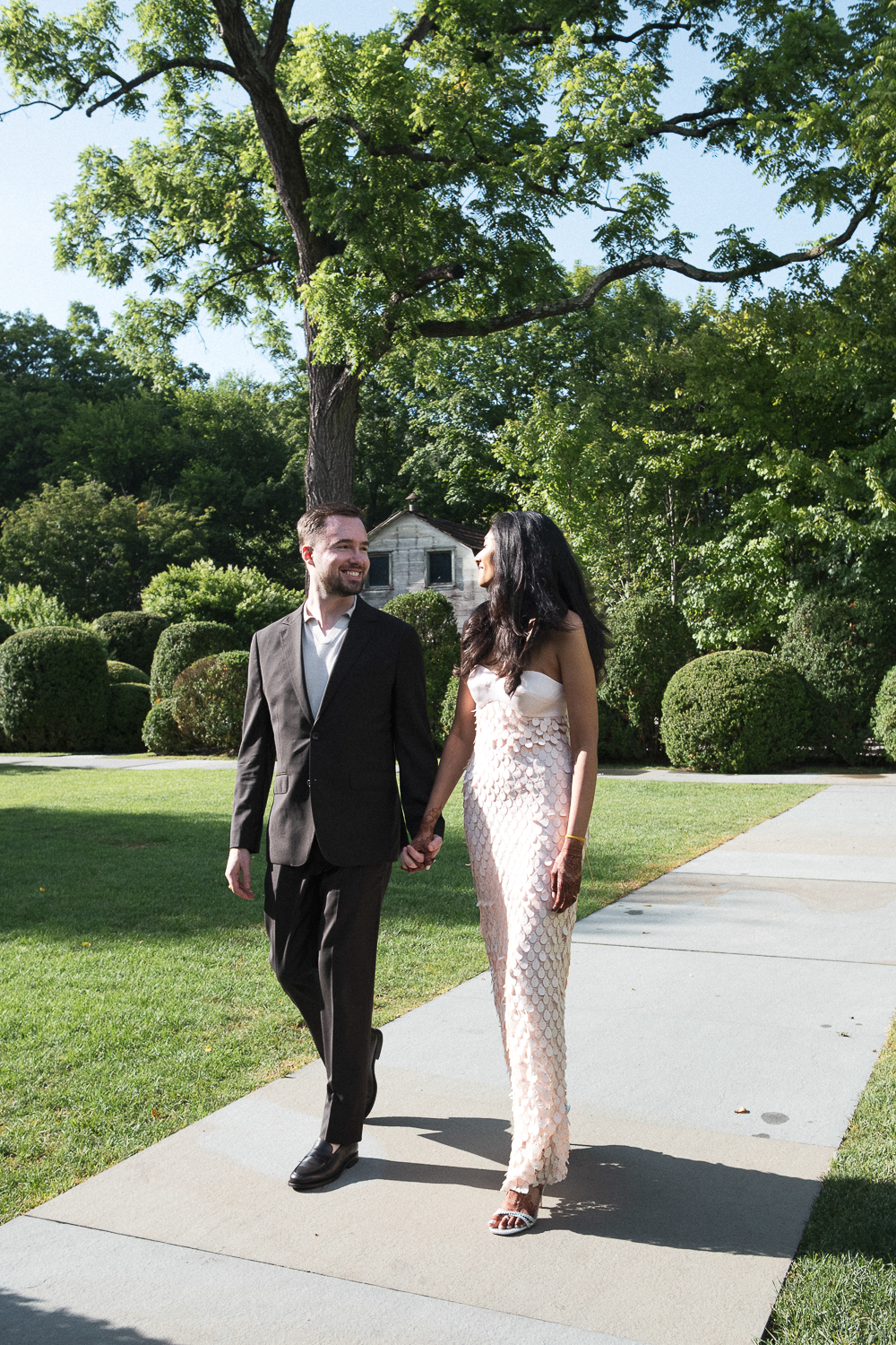 A couple walking hand in hand on a sidewalk in a park with lush green trees and bushes on a sunny day.