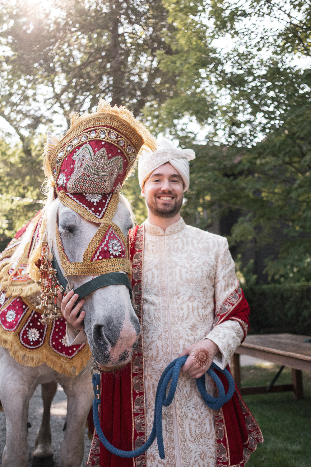 A man dressed in traditional South Asian bridal attire standing beside a white decorated horse outdoors during daytime.