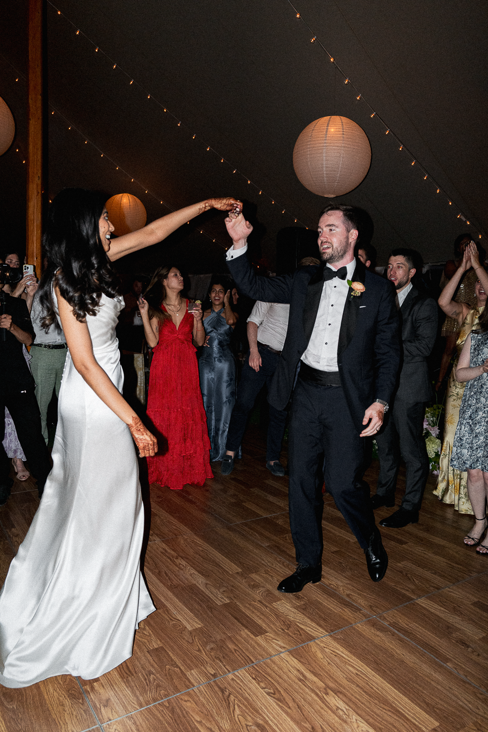 A bride and groom dancing at their wedding reception, surrounded by guests in formal attire, under hanging lantern lights.
