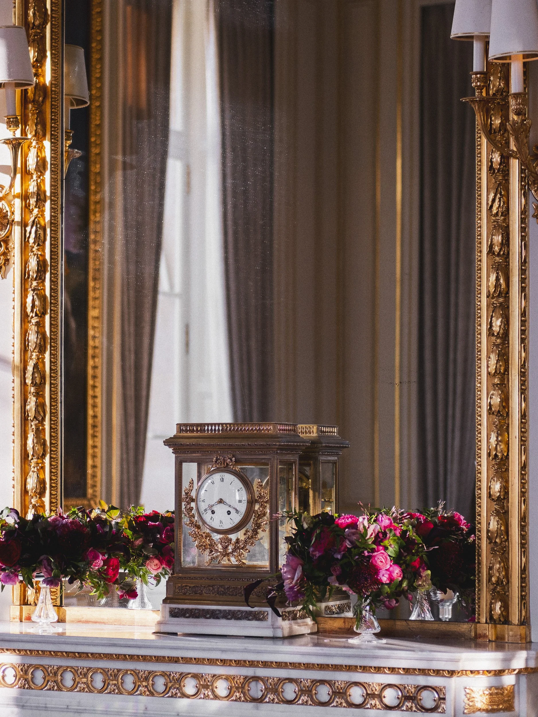 An ornate gold-framed mirror reflects a room with a marble-topped table decorated with pink flowers and an antique clock. The reflection shows drapes and part of the ceiling with gold moldings.