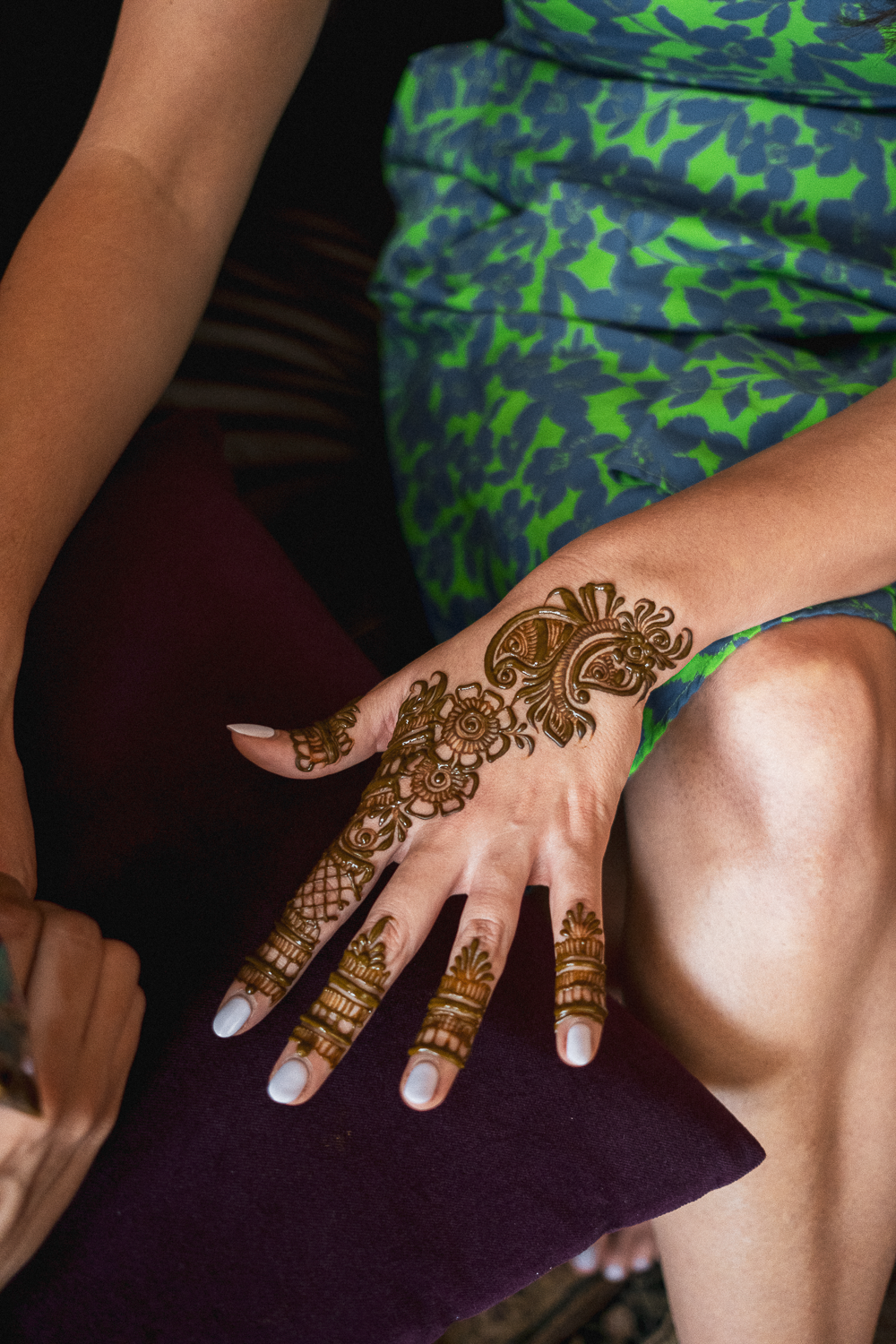 A woman with intricate henna designs on her hand and fingers, wearing a blue and green patterned dress, sitting on a maroon cushion.